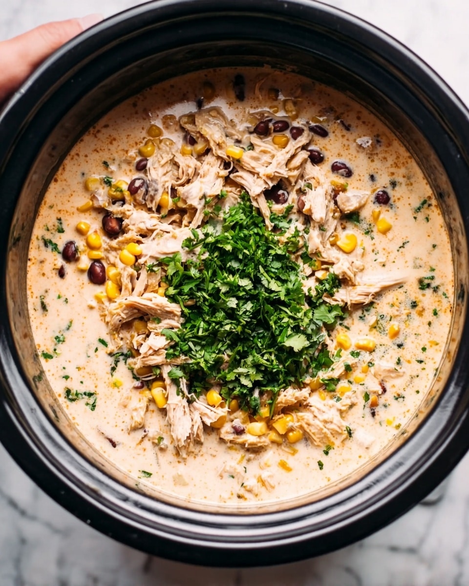 The image shows a large slow cooker filled with a creamy soup base that is light beige in color, spread evenly at the bottom and around the edges. Shredded light brown chicken pieces are scattered on top throughout the soup, interspersed with dark black beans and small yellow corn kernels. In the center, there is a generous mound of finely chopped bright green herbs, likely cilantro or parsley, adding a fresh contrast to the creamy and chunky mixture. The slow cooker sits on a white marbled surface. A woman's hand is holding the slow cooker from the side. Photo taken with an iphone --ar 4:5 --v 7
