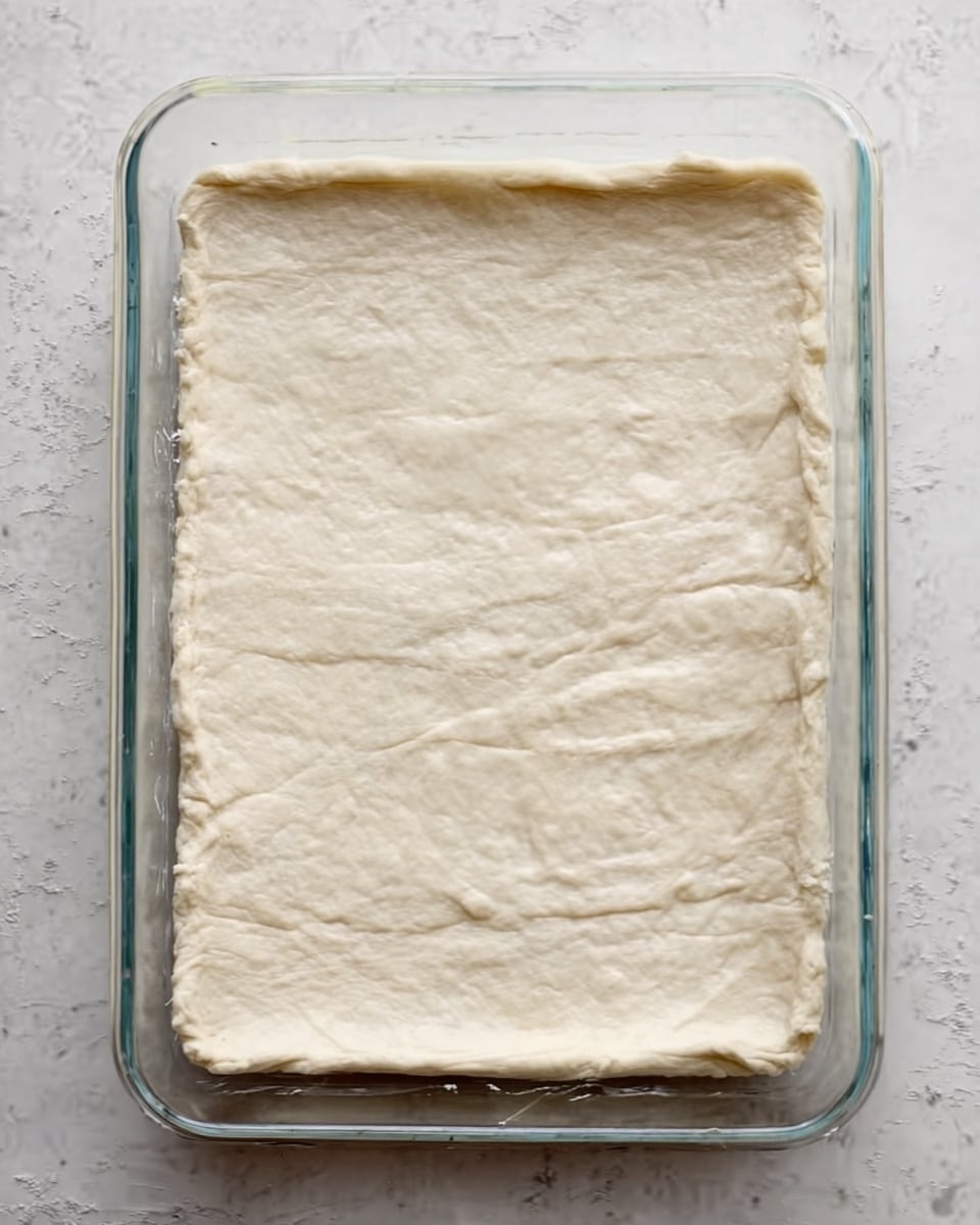 A rectangular glass baking dish filled with a single layer of soft, pale dough that covers the entire bottom surface. The dough appears smooth but with light creases and slight folds near the edges, showing it is gently pressed to fit the shape of the dish. The edges are slightly thicker compared to the center, which looks even and flat. The dish sits on a white marbled textured surface. photo taken with an iphone --ar 4:5 --v 7