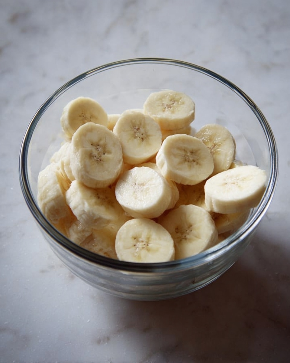Magnolia Bakery's Famous Banana Pudding Recipe 4 The image shows a clear glass bowl filled with round slices of banana stacked inside. The bowl sits on a white marbled surface that brightens the background. The banana slices are pale yellow with a smooth texture, arranged loosely without neat layering. Light shines softly on the bowl, making the glass and bananas clear and fresh-looking. Photo taken with an iphone --ar 4:5 --v 7