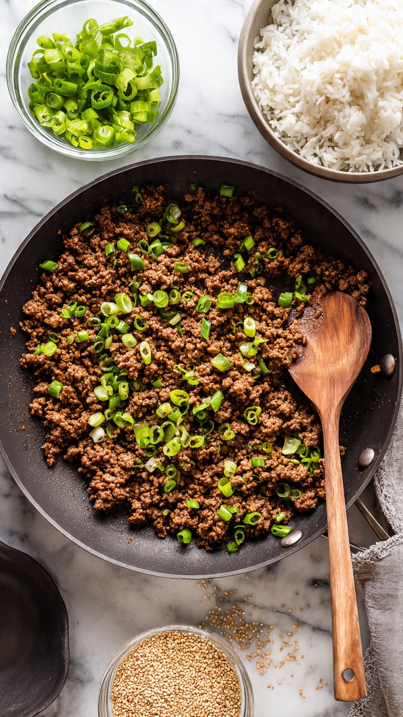 A close-up top view of a dark pan filled with cooked ground meat browned with small green onion pieces scattered on top. A wooden spoon with a light handle rests inside the pan on the right side. Above the pan, there is a bowl of white rice, and to the left of the rice is a small clear bowl filled with sliced green onions. Below the pan, a small glass bowl holds golden sesame seeds with some seeds scattered around it on a white marbled surface. A dark gray pan sits empty in the lower left corner. photo taken with an iphone --ar 4:5 --v 7