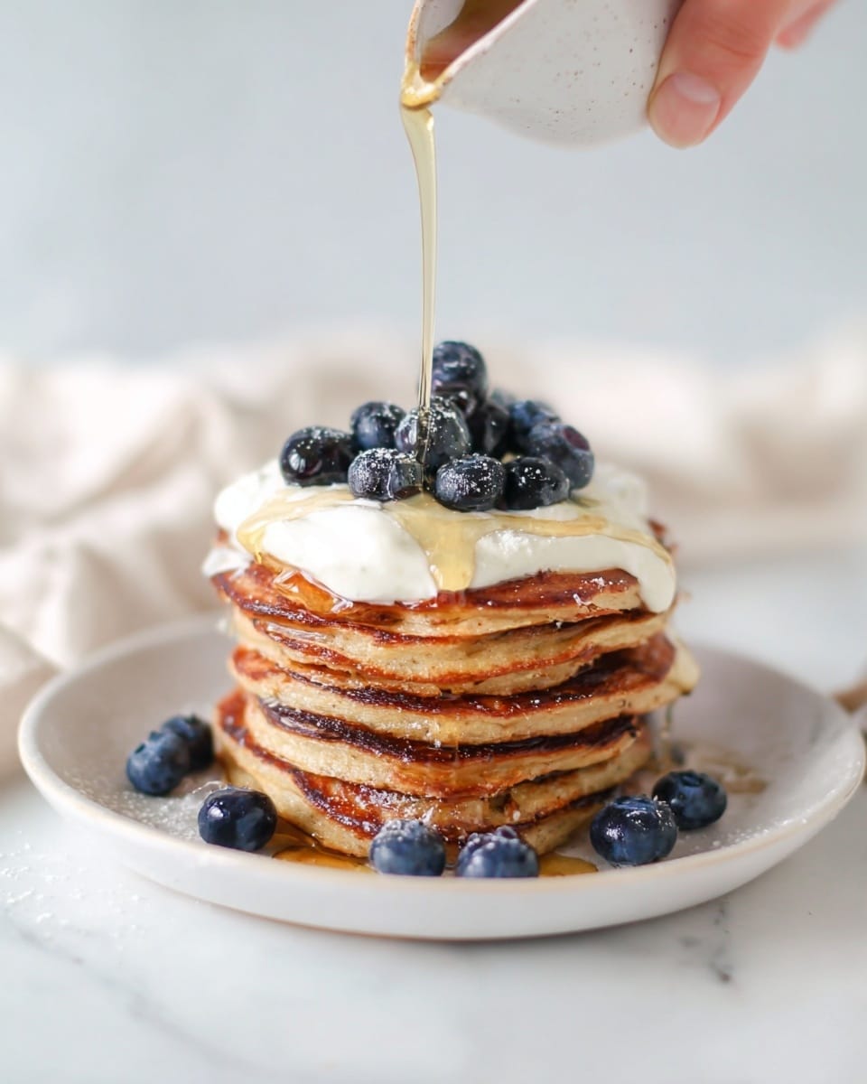A stack of six golden brown pancakes is placed on a white plate sitting on a white marbled surface. The pancakes have a slightly crispy edge and a soft texture. On top, there is a layer of white cream with fresh dark blue blueberries scattered around and on top of the cream. More blueberries are placed at the base of the pancake stack on the plate. A woman's hand is pouring clear golden syrup over the stack, with syrup streaming down the sides of the pancakes. The scene is bright and simple, with a soft background photo taken with an iphone --ar 4:5 --v 7