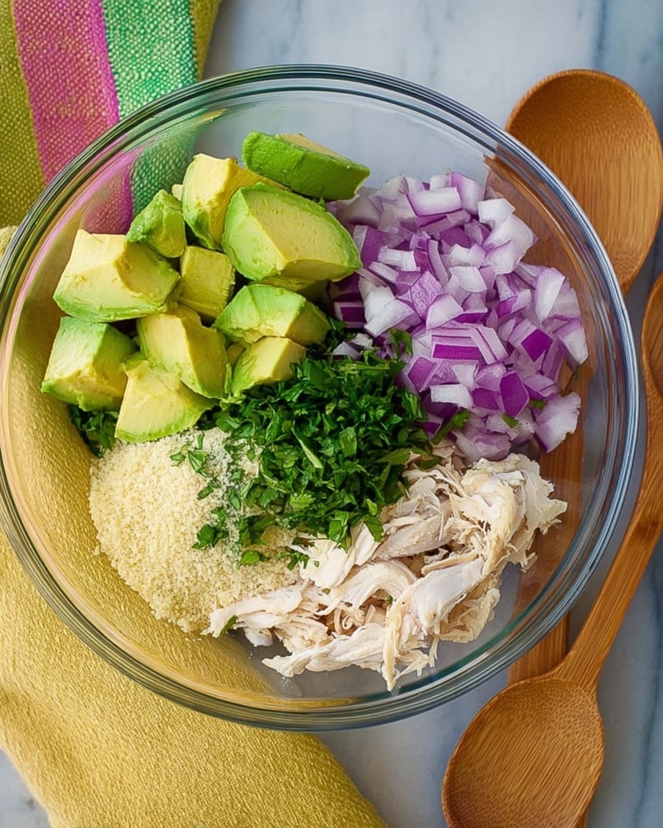 A clear glass bowl placed on a white marbled surface contains five visible layers of ingredients arranged in sections. Starting at the top left, there are chunks of bright green avocado with a smooth texture. Next to the avocado on the right is a pile of chopped purple-red onion with a firm texture. Below the onion is shredded white chicken with a soft, fibrous texture. To the left of the chicken is chopped fresh green cilantro with a leafy texture. Below the cilantro is a small pile of light beige breadcrumbs or similar small crumbs. To the right of the bowl on the white marbled surface, there are two wooden spoons and a yellow cloth with green and pink stripes. Photo taken with an iphone --ar 4:5 --v 7
