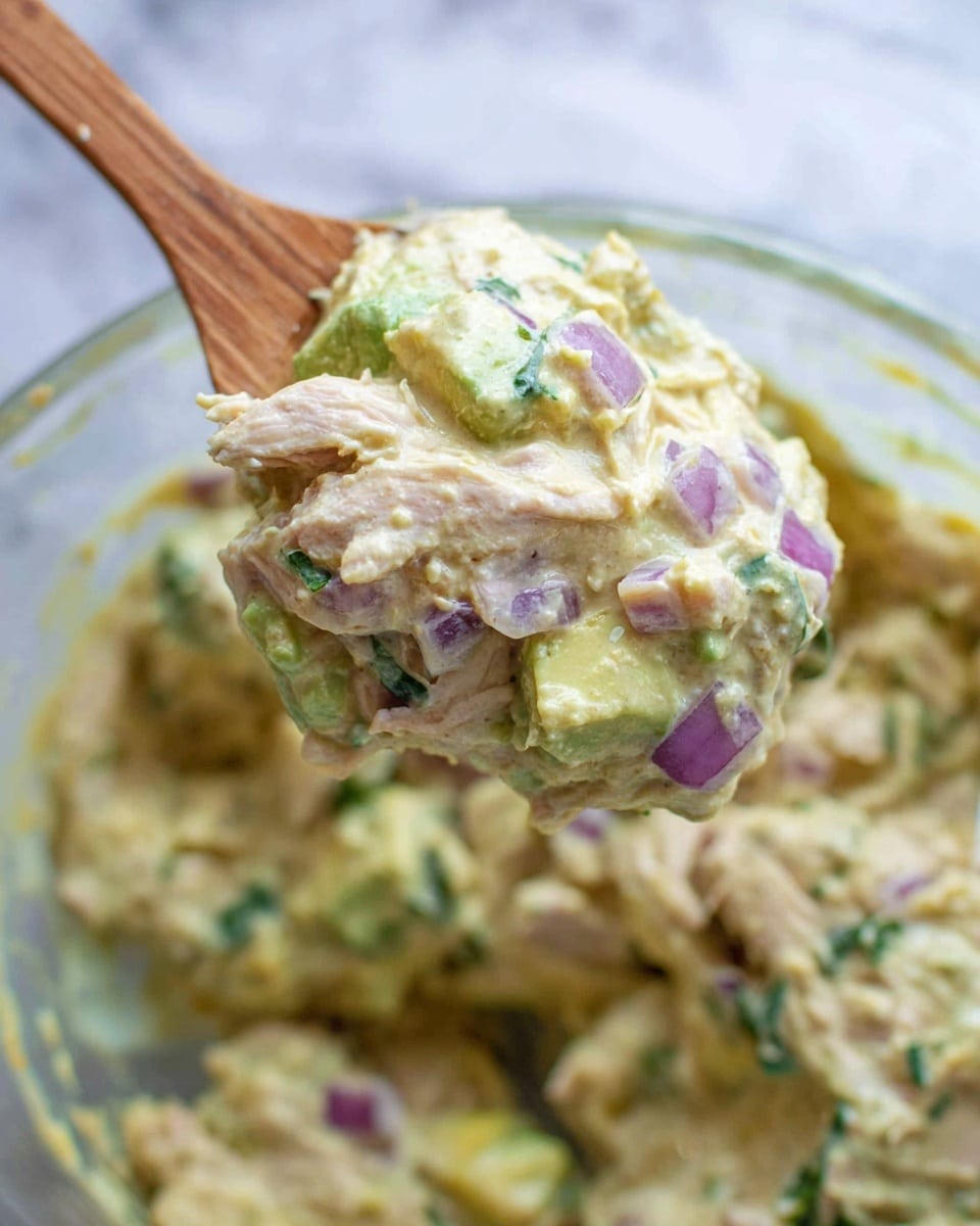 The image shows a creamy mixture held by a wooden spoon above a clear bowl, filled with a chunky blend of shredded pale meat, green avocado pieces, small purple onion cubes, and finely chopped green herbs, all mixed in a thick, light yellowish sauce with a slightly lumpy texture. The background is a white marbled texture, giving a clean and fresh look. photo taken with an iphone --ar 4:5 --v 7
