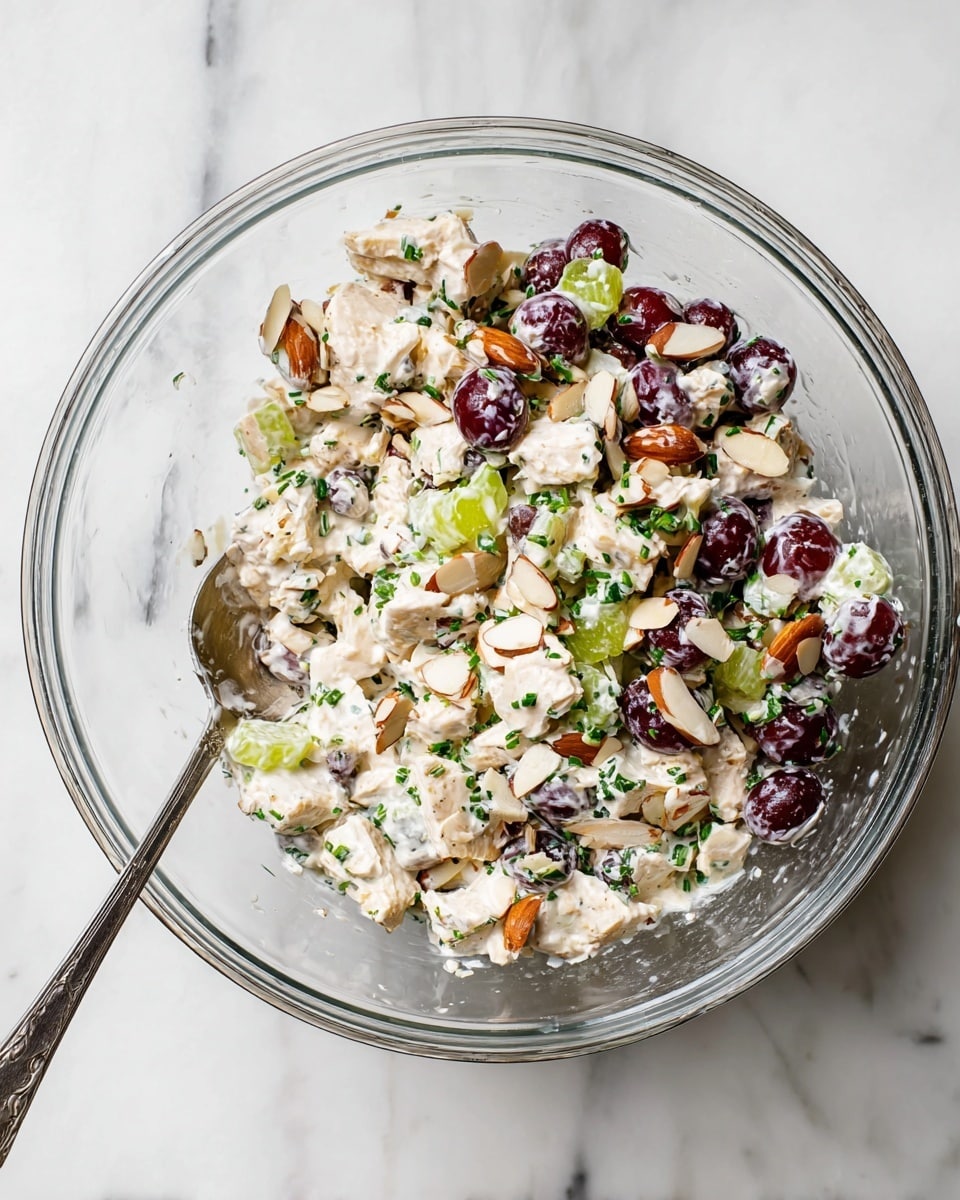 A clear glass bowl filled with a mixed salad that has three main layers: the bottom is a creamy white dressing, the middle layer shows small light beige chunks of cooked chicken, and the top layer has scattered pieces of dark red grapes cut in halves, chopped green celery, sliced almonds in light brown, and small green herbs finely chopped and mixed through the salad. A silver spoon is partially placed inside the bowl on the left side. The bowl sits on a white marbled surface. photo taken with an iphone --ar 4:5 --v 7
