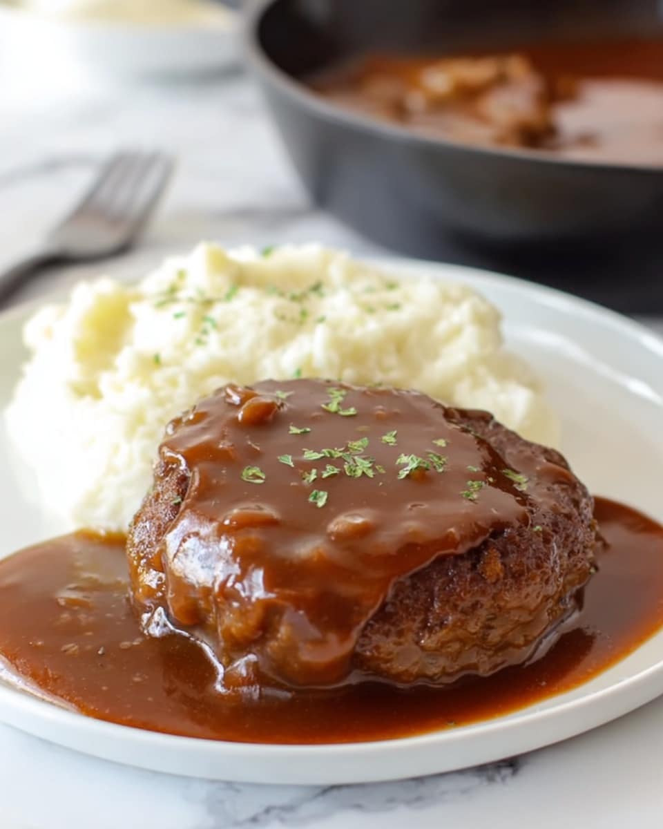 The image shows a round meat patty covered with a thick, dark brown gravy that looks smooth and shiny, sitting on a white plate. The gravy has small green herbs sprinkled on top, adding a touch of color. To the left of the meat patty, there is a scoop of white mashed potatoes with a soft and fluffy texture. The plate is on a white marbled surface, and in the background, there is a black pan partially visible with more gravy inside. Photo taken with an iphone --ar 4:5 --v 7