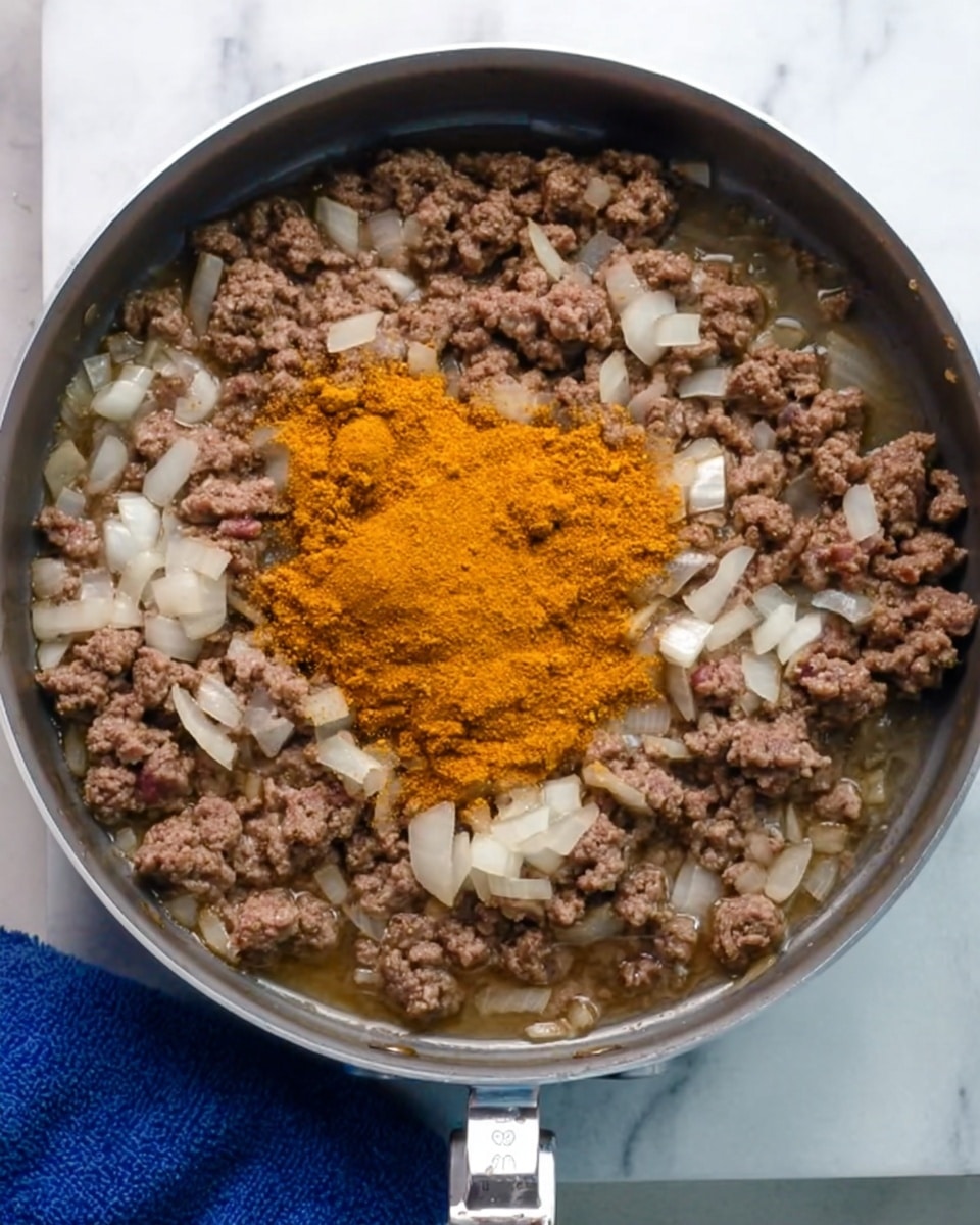 A top view of a round frying pan on a white marbled surface, filled with cooked ground meat and chopped onions. In the center of the pan, there is a layer of orange-yellow powdery spice resting on top of the meat mixture. The meat is brown with some pink spots, and the onions are translucent white, spread evenly throughout the pan. The pan's handle extends to the right side, and part of a blue cloth is visible on the left side under the pan. photo taken with an iphone --ar 4:5 --v 7