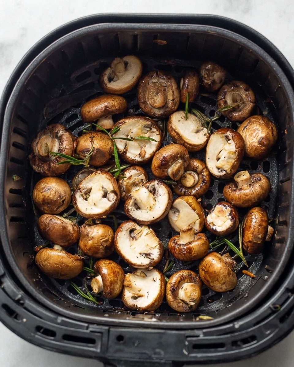 Inside a black air fryer basket, there are many whole and sliced brown mushrooms scattered evenly, showing a mix of light brown and creamy white colors with a slightly cooked texture. Small green rosemary sprigs are visible among the mushrooms, adding a touch of color contrast. The basket's ridged surface and the mushrooms' soft, slightly browned edges create a textured, home-cooked look, all placed on a white marbled background. photo taken with an iphone --ar 4:5 --v 7