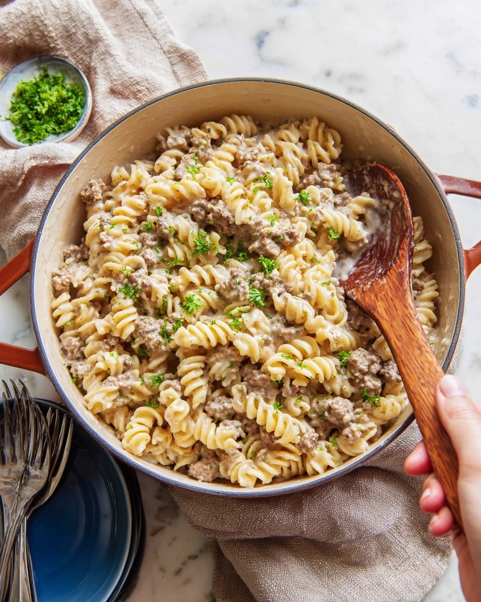 A large white pot filled with creamy pasta mixed with ground meat, showing about two layers of spiraled noodles coated in a light sauce. Some small green parsley leaves are scattered on top, adding a fresh touch. A woman's hand holds a wooden spoon stirring the pasta on the right side. The pot sits on a white marbled surface with a beige cloth partly under it and some blue bowls with silver forks in the background. Photo taken with an iphone --ar 4:5 --v 7