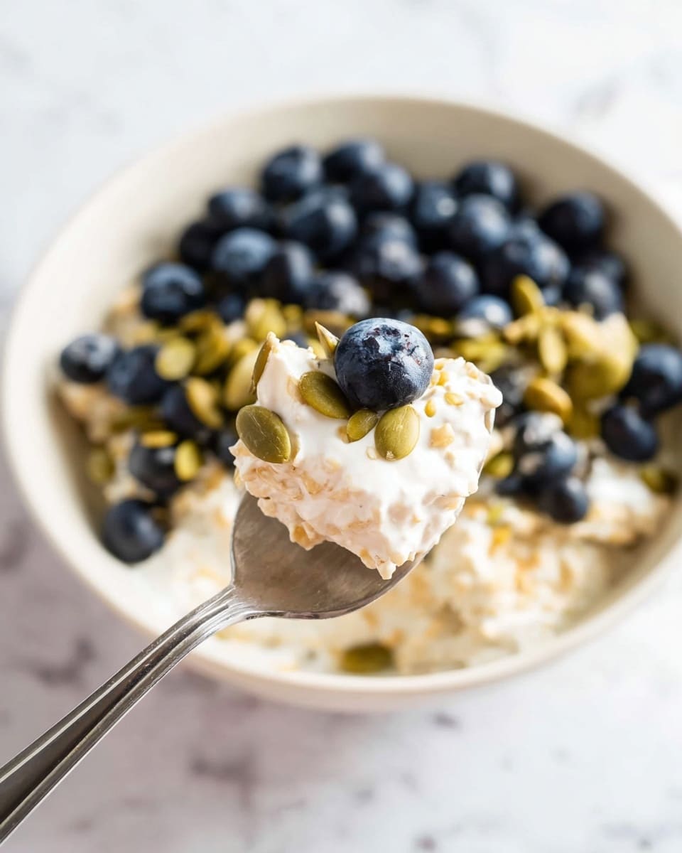 A white bowl holds a three-layer dish starting with a creamy white oatmeal base at the bottom. On top of this is a layer of green pumpkin seeds and golden oats, giving a crunchy texture. The top layer consists of dark blue round blueberries spread evenly across. A spoon with a shiny silver surface is shown close up in the foreground holding a scoop of oatmeal with a blueberry and pumpkin seeds resting on it. The background has a soft white marbled texture. photo taken with an iphone --ar 4:5 --v 7