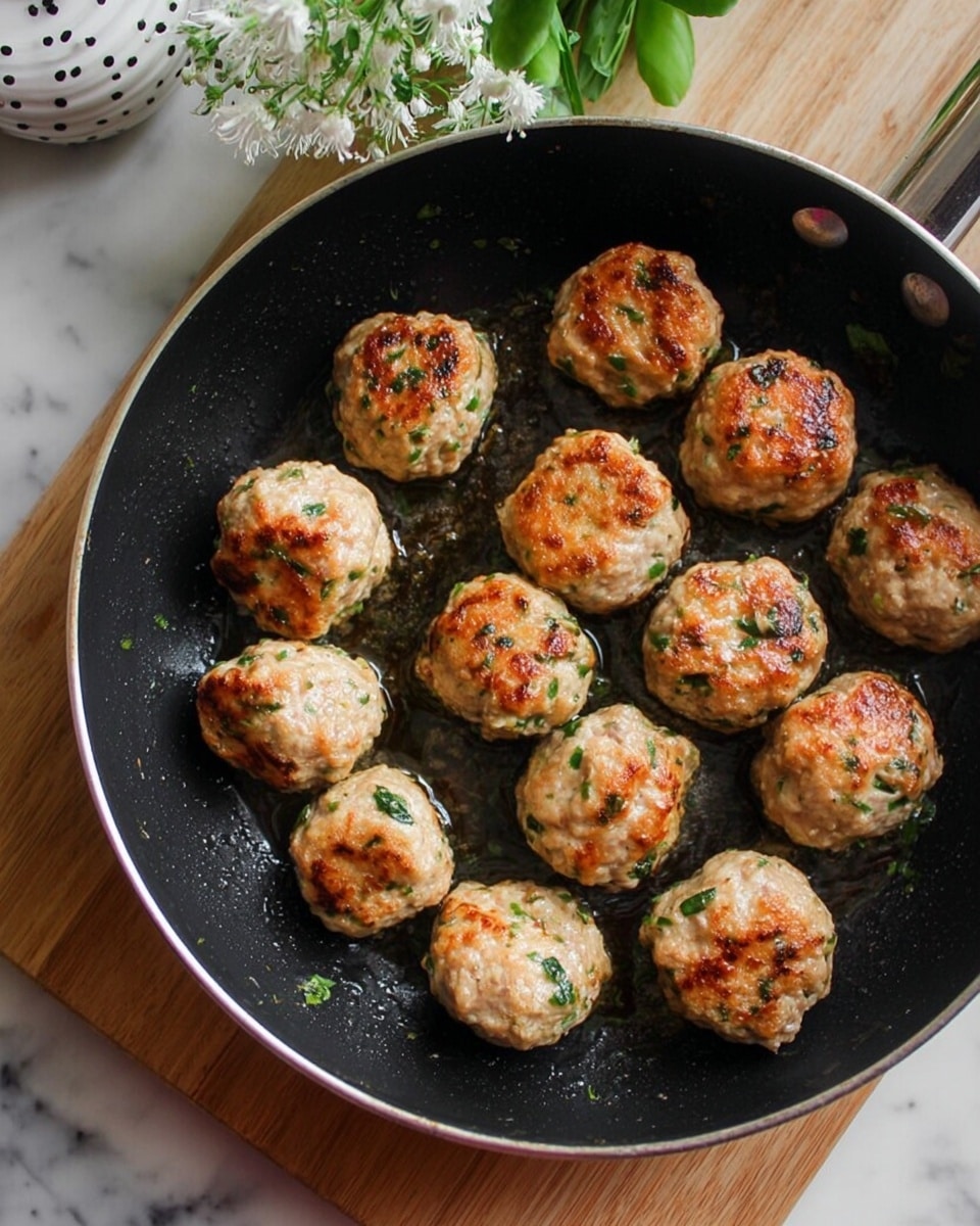 The image shows a black frying pan placed on a wooden board with twelve browned meatballs inside it. Each meatball is light brown with visible green herbs and has a slightly crispy texture on the outside. They are arranged closely but not touching much, and the pan has a bit of oil giving a shiny look. In the top part of the image, a small bunch of white flowers with green leaves and a white cup with black dots are partly visible. The whole scene is set on a white marbled surface. Photo taken with an iphone --ar 4:5 --v 7