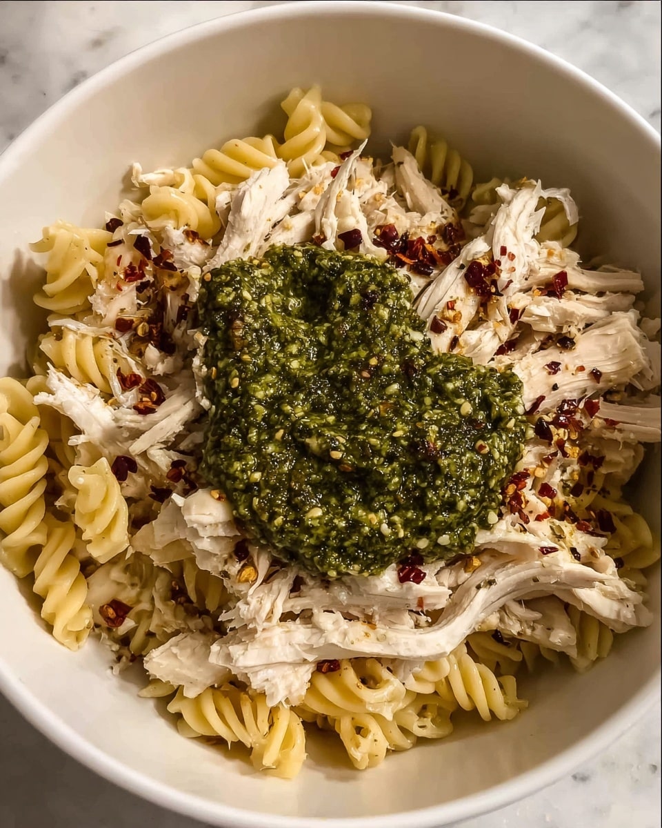 A white bowl holds three clear layers: at the bottom, light yellow rotini pasta fills the space with its spiral shapes; above it, shredded white chicken is spread all over, sprinkled lightly with red chili flakes and black pepper; on top, a dollop of thick dark green pesto sauce sits in the center, adding a textured contrast to the pale ingredients below. The background is a white marbled surface. photo taken with an iphone --ar 4:5 --v 7