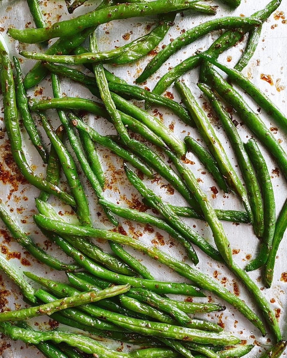 The image shows many roasted green beans spread out on a slightly browned baking sheet. The beans are bright green with a slight shine from oil, some wrinkles on the surface, and small dark spots from roasting. The beans are arranged unevenly across the silver, textured baking sheet with scattered tiny brown crisp bits around them. The background is changed to a white marbled texture. Photo taken with an iphone --ar 4:5 --v 7