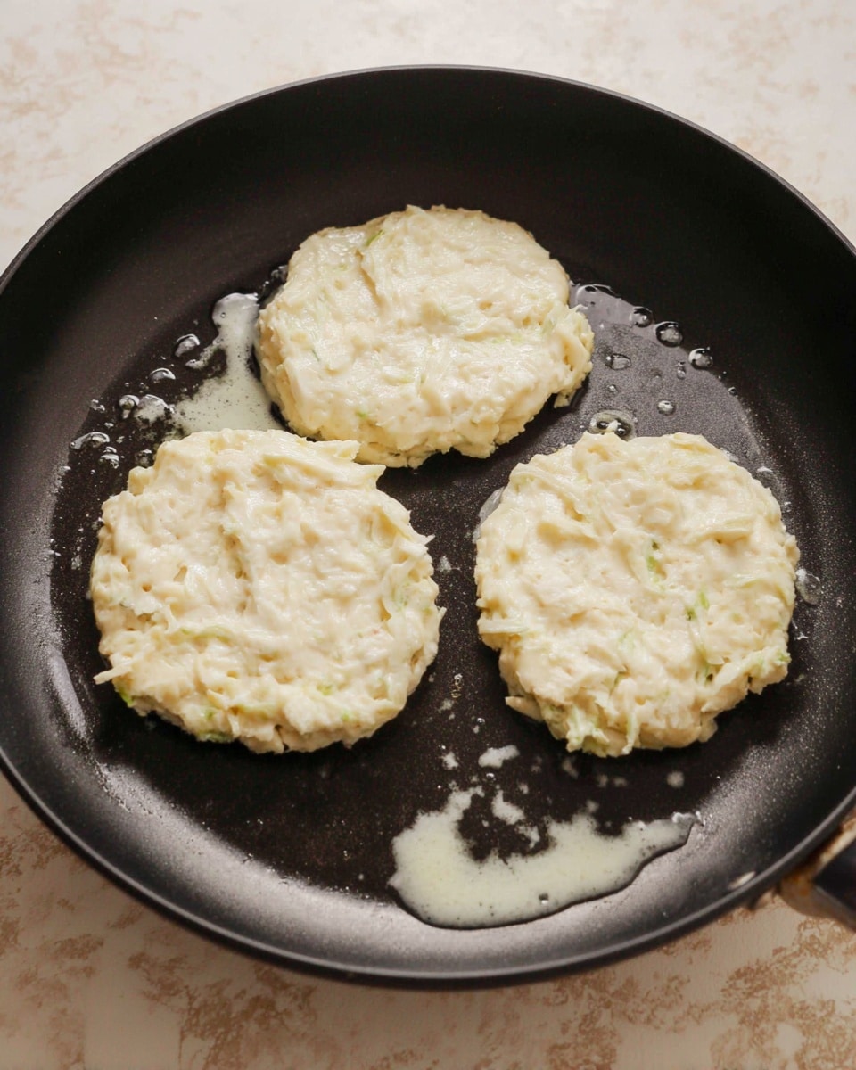 Three white, soft, and slightly rough-textured patties cook in a black pan with a thin layer of oil. Each patty looks uneven with small chunks and strands, giving a fresh, moist appearance. The pan sits on a white marbled surface. The background shows a bit of a beige counter. Photo taken with an iphone --ar 4:5 --v 7