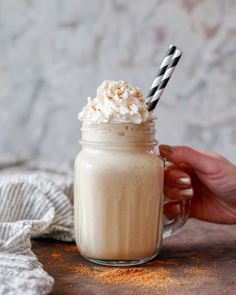 The image shows a creamy light beige drink in a clear glass jar with a handle, filled almost to the top. On top, there is a large swirl of white whipped cream sprinkled with light brown powder. A black and white striped straw sticks out from the whipped cream. A woman's hand with natural nails is holding the jar handle gently. The jar is placed on a brown wooden surface with scattered light brown powder, and a white and gray striped cloth is laid nearby. The background has a soft focus with a white marbled texture. photo taken with an iphone --ar 4:5 --v 7