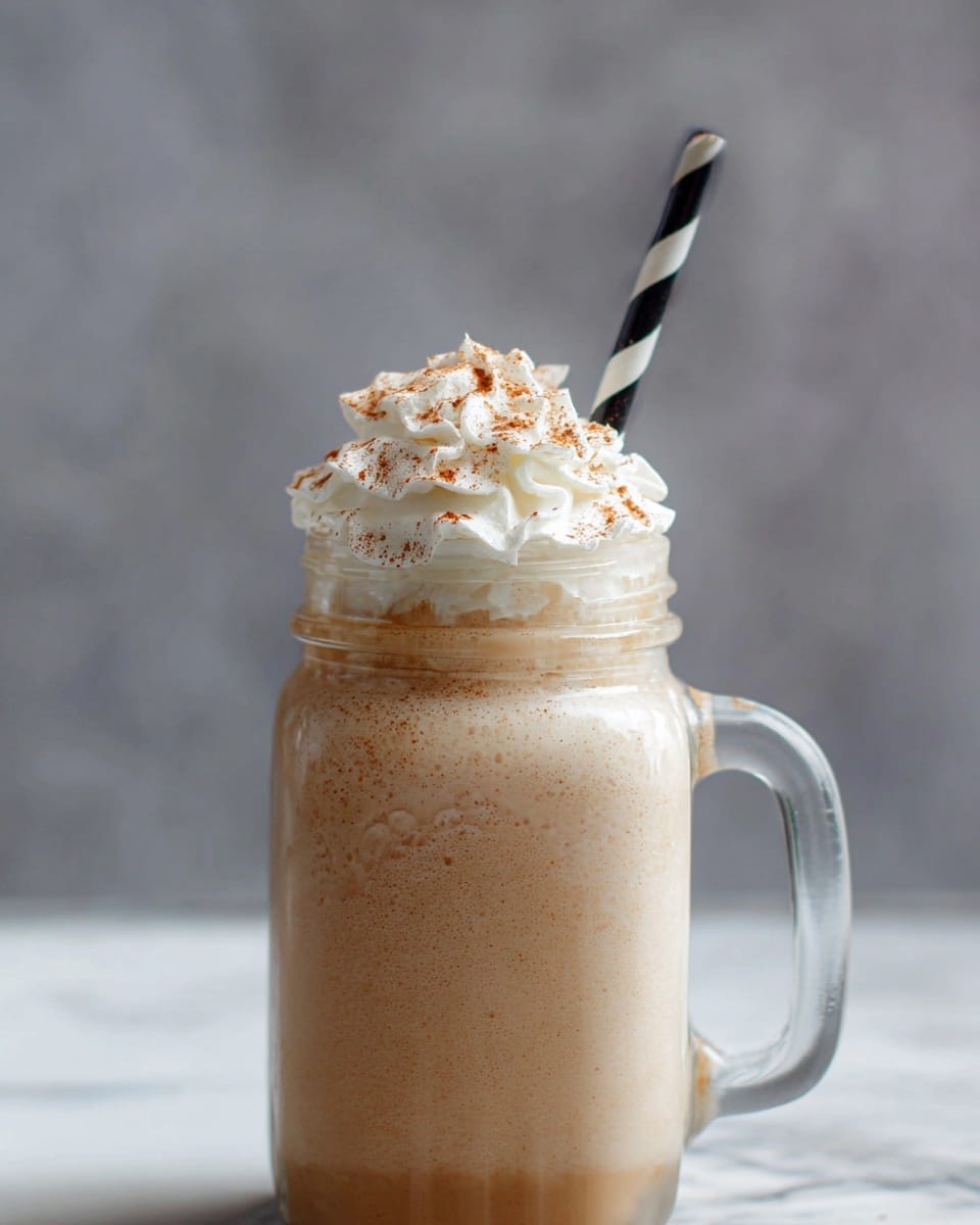 A tall clear glass jar with a handle is filled with a light brown creamy drink topped with a fluffy swirl of white whipped cream. The whipped cream has a sprinkle of brown powder on top, adding texture and color contrast. A black and white striped straw stands upright from the jar. The background is soft gray, and the jar sits on a white marbled surface. photo taken with an iphone --ar 4:5 --v 7