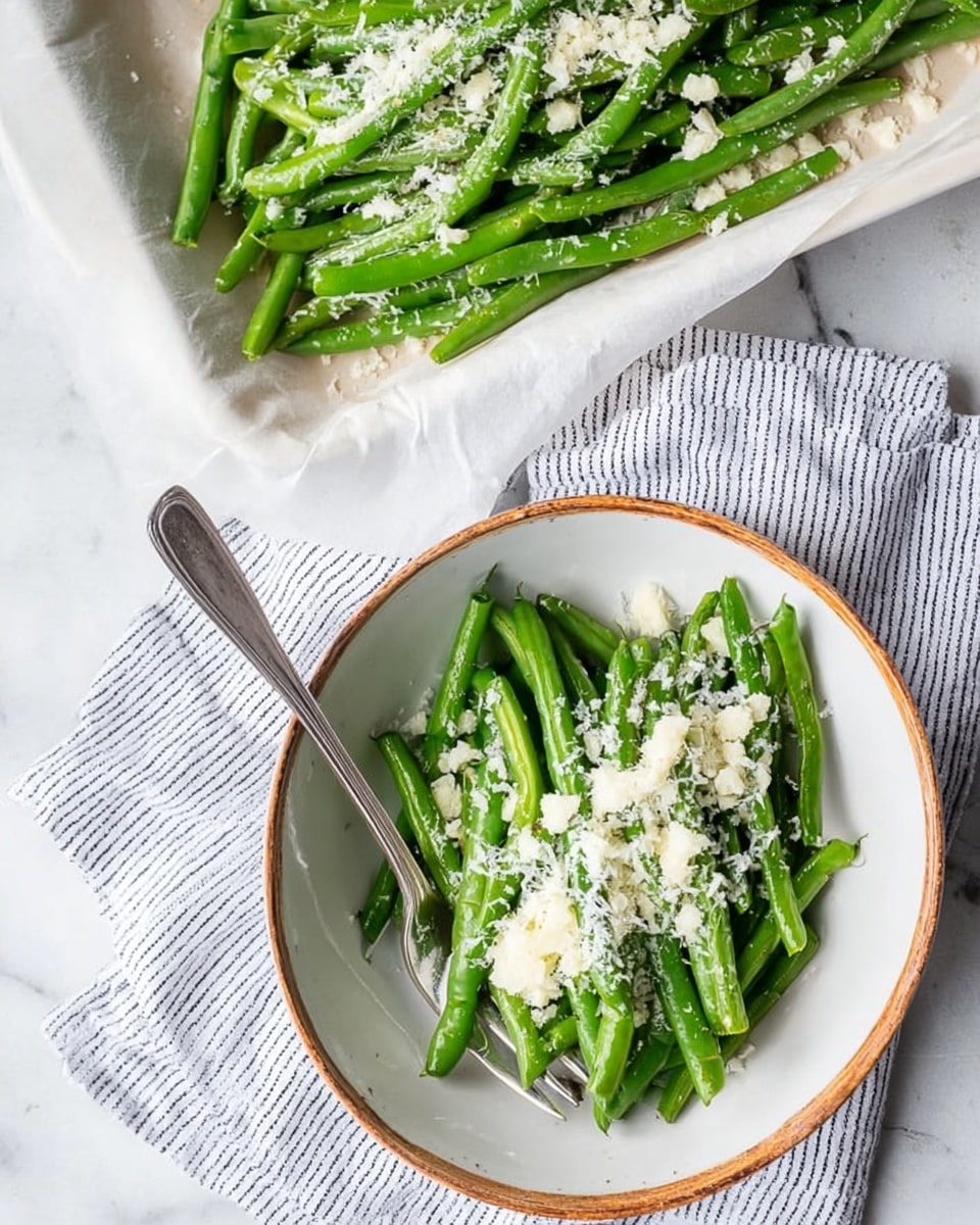 The image shows a white bowl filled with fresh green beans that are bright and shiny, some with light grill marks. On top of the green beans, there is a layer of finely grated white cheese scattered unevenly. A vintage silver fork rests inside the bowl on the left side, partially under the green beans. The bowl is placed on a white marbled surface with a striped cloth underneath on the bottom left corner. Photo taken with an iphone --ar 4:5 --v 7