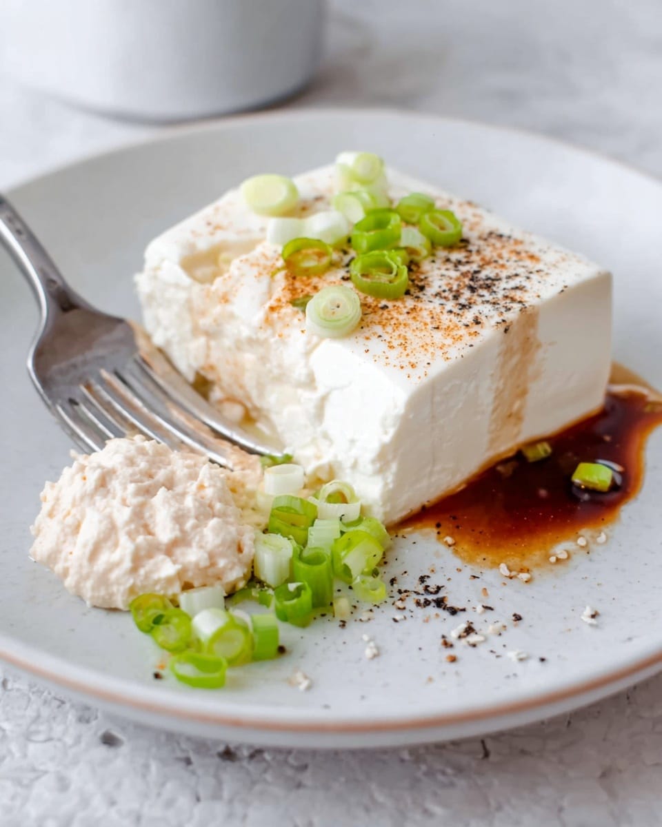 The image shows a white plate on a white marbled surface. On the plate is a soft, white tofu block lightly dusted with a brown powder on top. Around the tofu, there is a dark brown sauce on one side, black pepper sprinkled near the tofu, and a small pile of chopped green onions stacked beside it. Next to the tofu, there is a dollop of light pink paste with visible soft texture. A silver fork is placed on the left edge of the plate, touching the dollop. Photo taken with an iphone --ar 4:5 --v 7