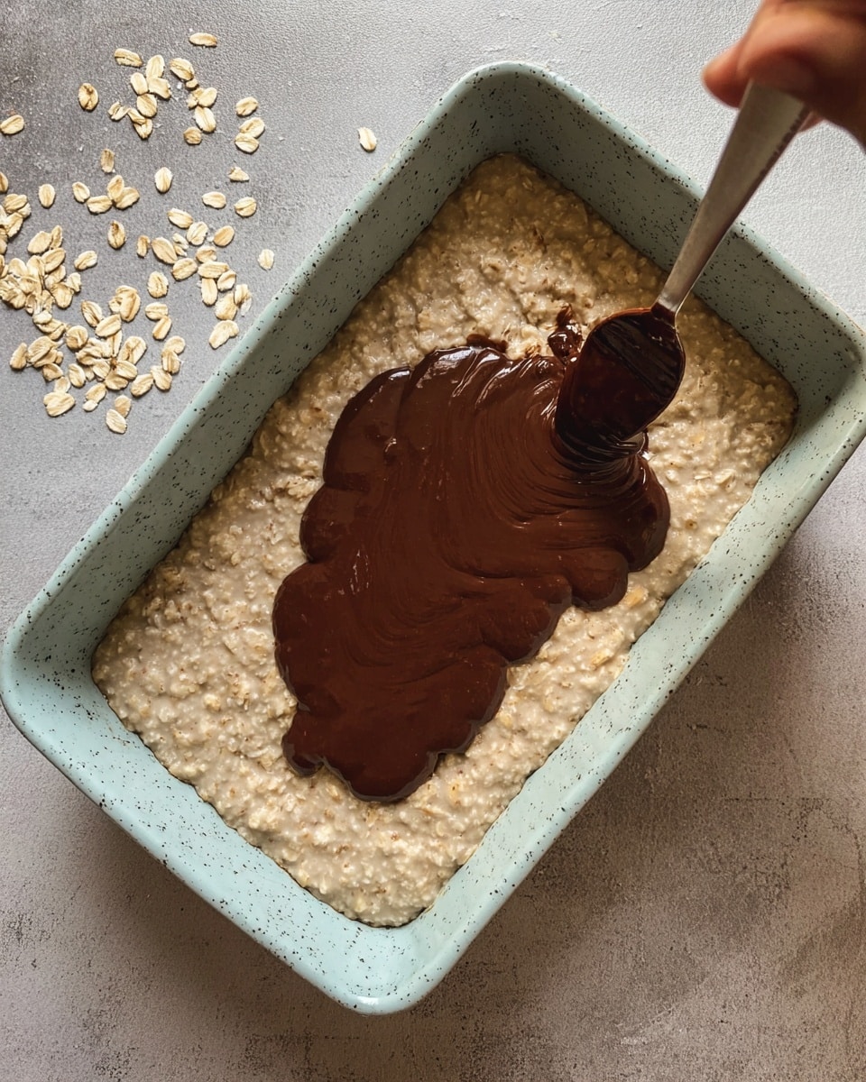 The image shows a light blue speckled rectangular baking dish filled with a beige, thick oatmeal base as the first layer. A dark brown chocolate sauce is being spread over the oatmeal in the center, covering about half the surface in an uneven shape. A woman’s hand holding a spoon with some chocolate sauce is visible on the right side, just above the dish. The dish is placed on a neutral grey surface with some scattered oats around. The oatmeal has a grainy texture, while the chocolate sauce is smooth and glossy. photo taken with an iphone --ar 4:5 --v 7