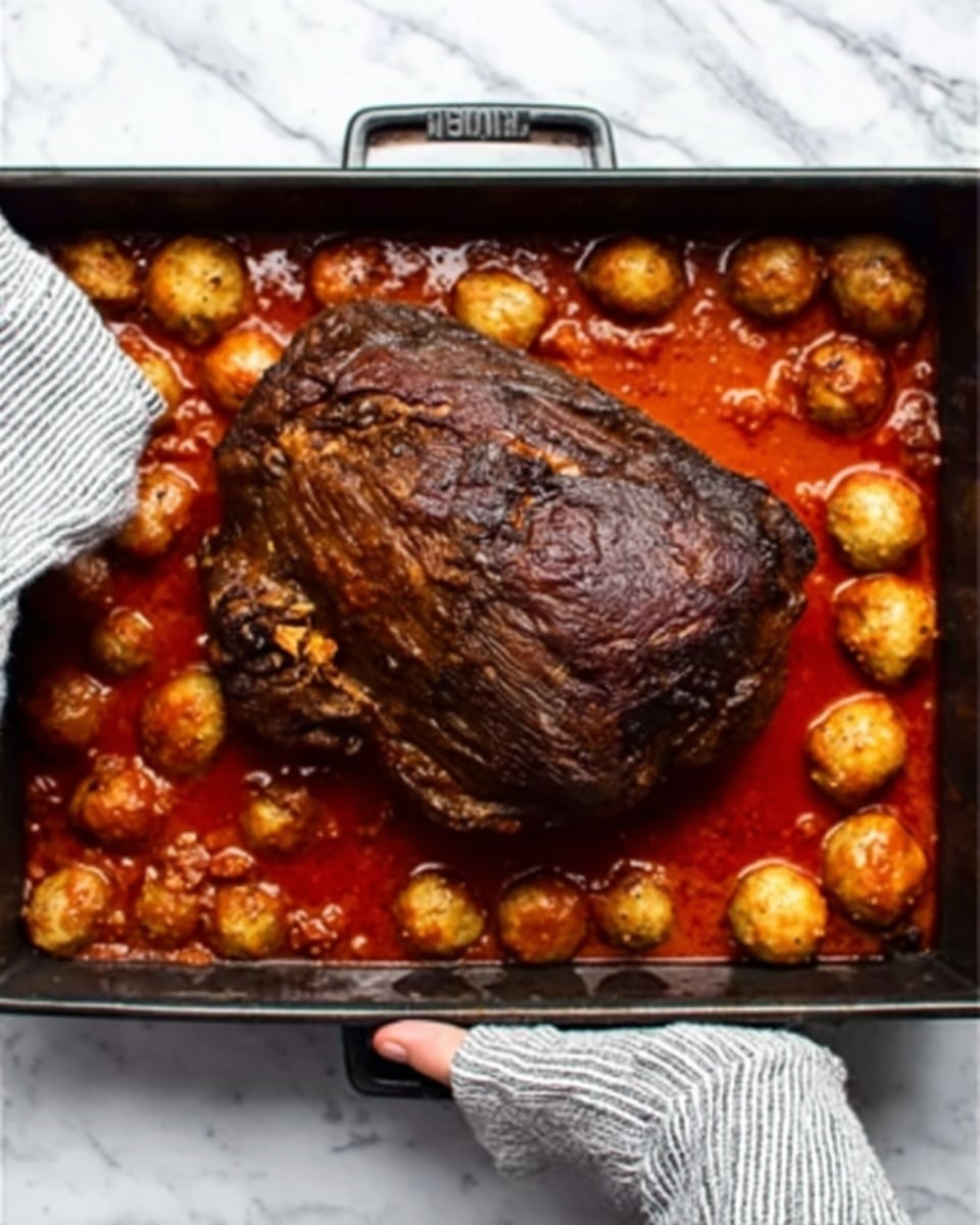 A large piece of cooked meat with a dark brown crust sits in the center of a black roasting pan filled with bright red tomato sauce. Surrounding the meat are many small, round meatballs with a light brown color, partially covered by the sauce. A woman's hand wearing a striped oven mitt holds the pan on the right side. The pan is placed on a white marbled surface. photo taken with an iphone --ar 4:5 --v 7
