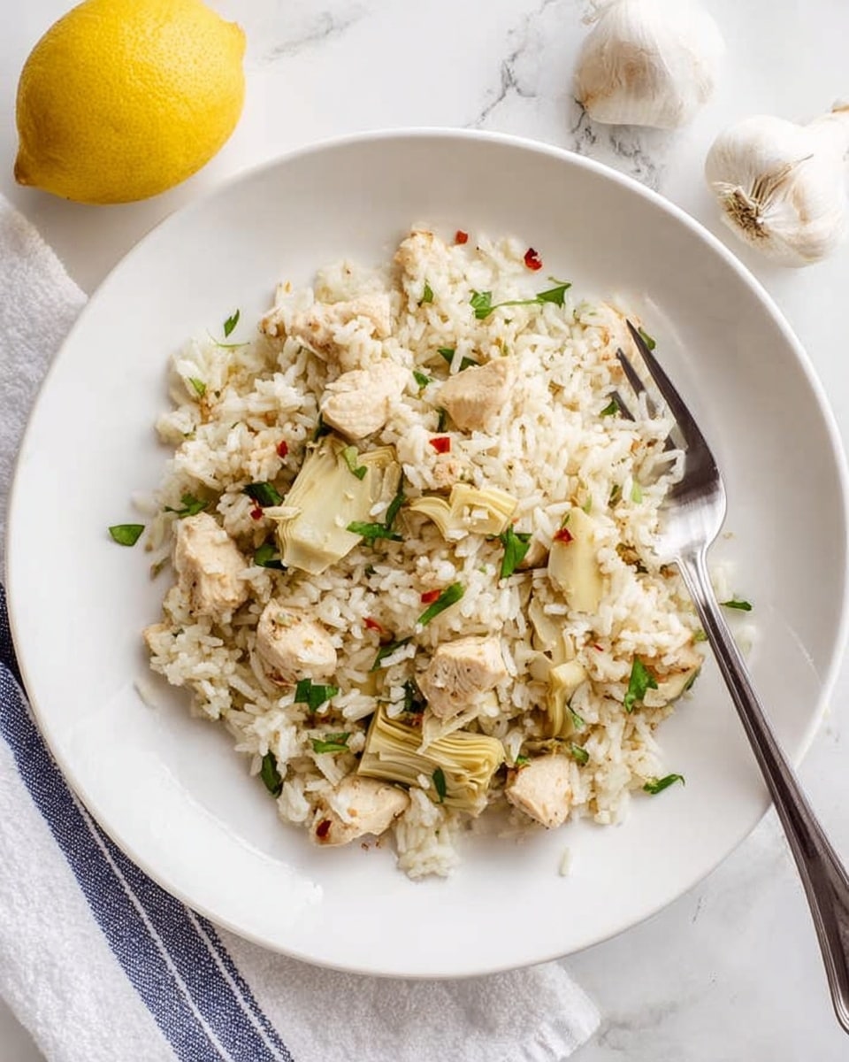 A white plate filled with a single layer of cooked white rice mixed with pieces of light beige chicken and pale yellow artichoke hearts scattered evenly throughout. The rice is fluffy with small bits of green herbs sprinkled on top, adding a touch of color, and some tiny red chili flakes. A silver fork rests on the right side of the plate, partly under the food. The plate sits on a white marbled surface, with a yellow lemon and a head of garlic positioned near the top left and right corners, and a white towel with blue stripes on the bottom left. Photo taken with an iphone --ar 4:5 --v 7