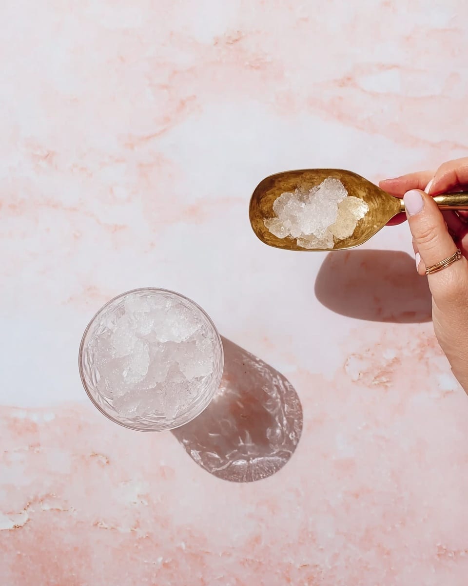 A woman's hand is holding a brass scoop filled with clear, crushed ice above a transparent glass that is empty on a white marbled surface. Next to it, there is another transparent glass partially filled with crushed ice. The background is a smooth white marble with soft pink veins. The scene is bright and clean. photo taken with an iphone --ar 4:5 --v 7