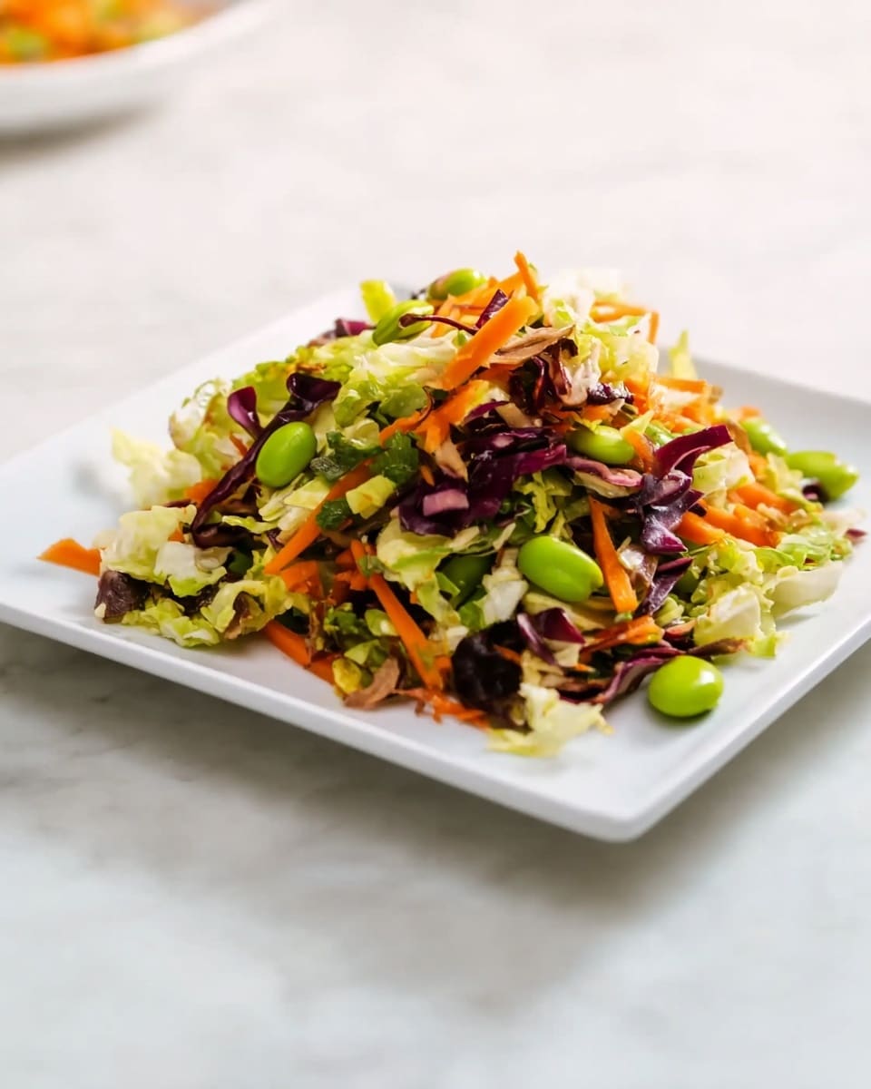 A white plate holds a colorful salad made of several layers: the bottom layer is finely chopped green cabbage, followed by thin strips of bright orange carrots and dark purple cabbage mixed evenly throughout. Scattered on top are green edamame beans adding a pop of brightness, and small bits of light green vegetables that add texture. The salad looks fresh and mixed well, sitting on a white marbled surface with a white cloth nearby. photo taken with an iphone --ar 4:5 --v 7