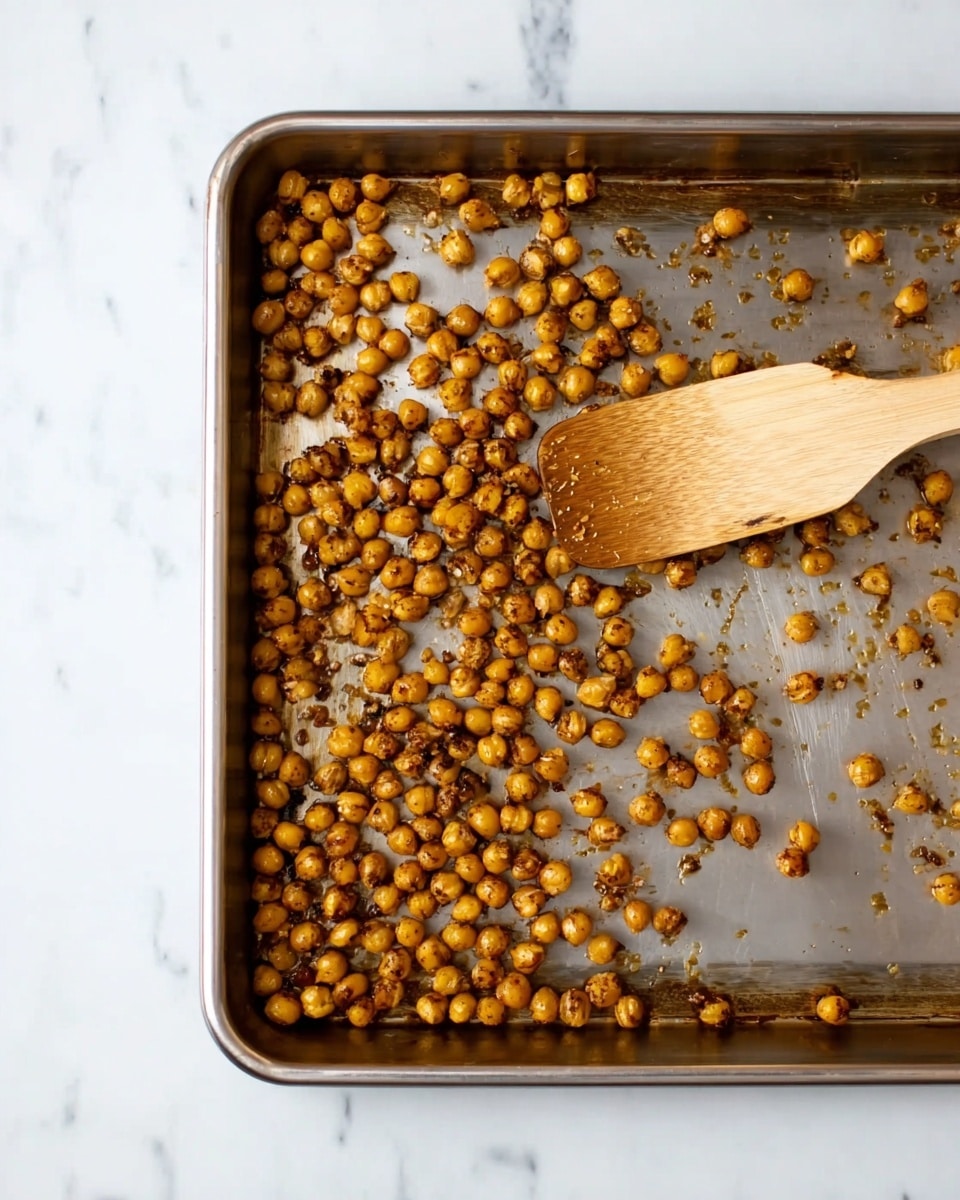 A metal baking tray filled with roasted chickpeas that are golden brown in color, scattered unevenly across the tray. A light wooden spatula rests inside the tray, positioned at the upper right corner, with the edge touching the chickpeas. The tray sits on a white marbled surface, showing a clean and simple kitchen setting. photo taken with an iphone --ar 4:5 --v 7