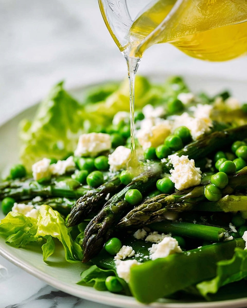 A fresh salad on a white plate shows layers of bright green lettuce leaves at the bottom, topped with green peas and dark green asparagus spears arranged evenly. White soft cheese is spread in small chunks across the salad, adding contrast to the green vegetables. A clear glass container is shown pouring a light yellow dressing over the top, catching the light and creating a glossy shine on the ingredients. The background is a white marbled surface that gives a clean, fresh look. photo taken with an iphone --ar 4:5 --v 7