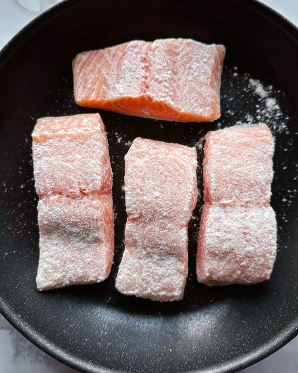 The image shows four raw salmon fillets in a black pan, each fillet dusted with a light layer of white flour. The salmon pieces are rectangular, arranged in two rows with two fillets in each row, showing a soft pink color with visible texture of the fish. The pan’s dark surface contrasts with the pale salmon and the white flour coating. The photo's overall setting includes a white marbled surface underneath the pan. photo taken with an iphone --ar 4:5 --v 7