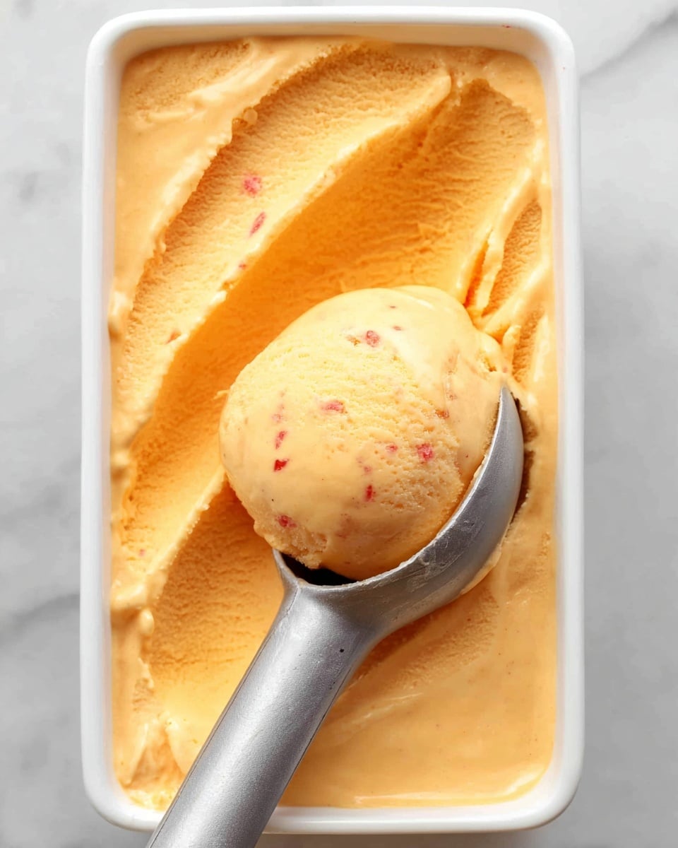 The image shows a close-up of a white rectangular dish filled with creamy orange ice cream with a smooth texture. The ice cream fills the dish fully and has a rounded scoop taken out near the center, held by a dull metal ice cream scoop. The scoop shows the same smooth orange ice cream with small red specks visible inside. The dish is placed on a white marbled surface with soft lighting highlighting the creamy texture of the ice cream. photo taken with an iphone --ar 4:5 --v 7