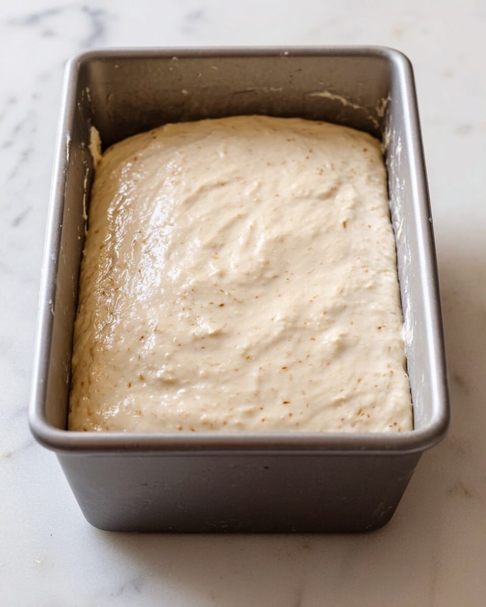 A metal baking pan filled with thick, light beige dough that has a creamy texture and is spread evenly to the edges, ready to be baked, sitting on a white marbled surface. The dough surface is smooth with some small air bubbles and faint brown specks throughout. Photo taken with an iphone --ar 4:5 --v 7