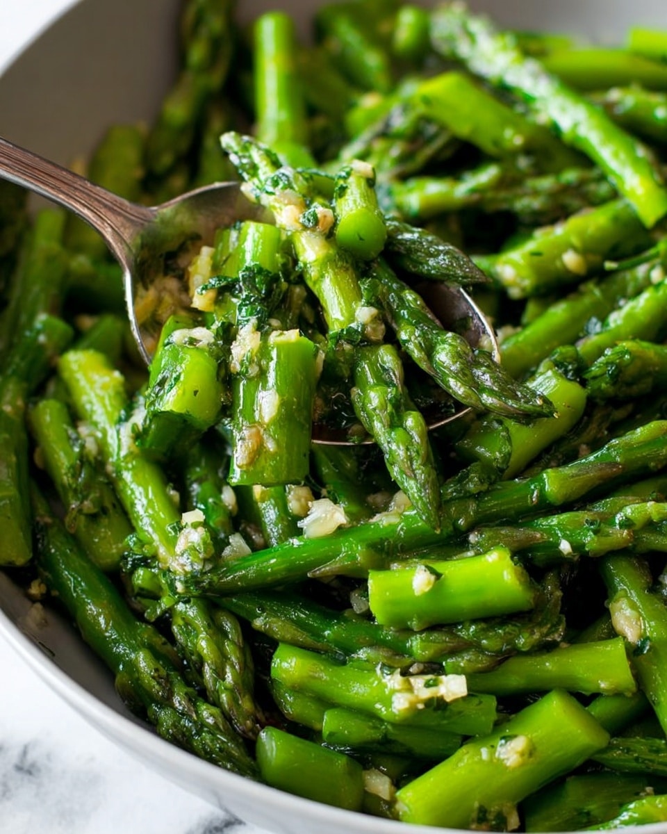 A close-up view of cooked asparagus in a white bowl, with many bright green asparagus pieces cut into short sections and mixed with small bits of garlic and herbs scattered all over. A silver spoon is scooping some asparagus near the center of the bowl. The asparagus looks glossy and seasoned, and the background is a white marbled texture. photo taken with an iphone --ar 4:5 --v 7