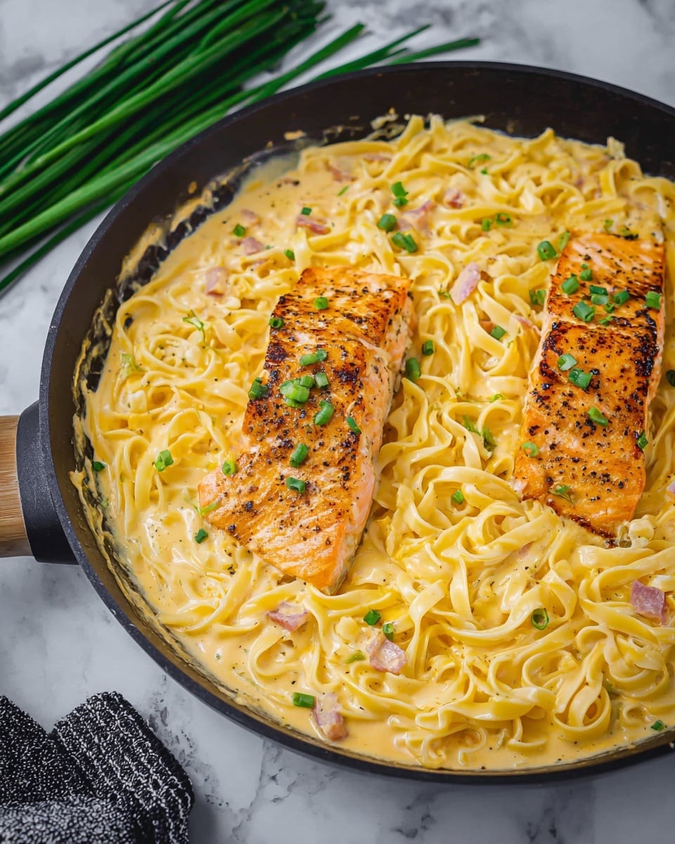 A close-up of a black pan filled with creamy yellow pasta sauce and fettuccine noodles evenly spread in the sauce, topped with two pieces of golden-brown cooked salmon sprinkled with small green chive pieces, all sitting on a white marbled surface with a bunch of fresh green chives on the left side and a black and white cloth near the bottom edge, photo taken with an iphone --ar 4:5 --v 7