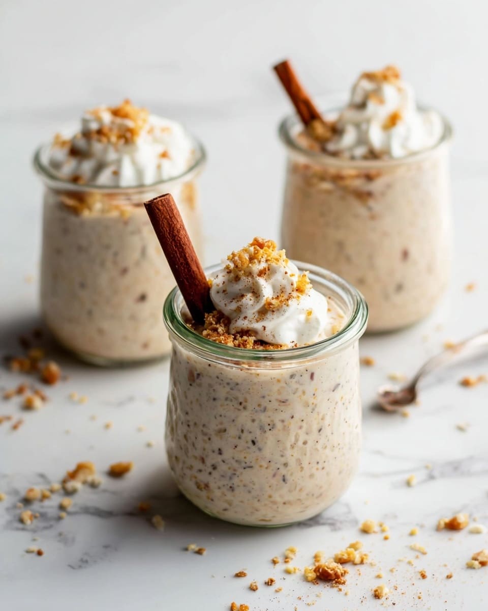 Three glass jars are filled with thick oatmeal overnight, light brown with visible oatmeal pieces. Each jar has a dollop of white cream on the top, sprinkled with crushed nuts that add a rough texture. Standing upright in the cream of each jar is a single dark brown cinnamon stick. The jars sit in a row on a white marbled surface with small bits of oats scattered nearby. photo taken with an iphone --ar 4:5 --v 7