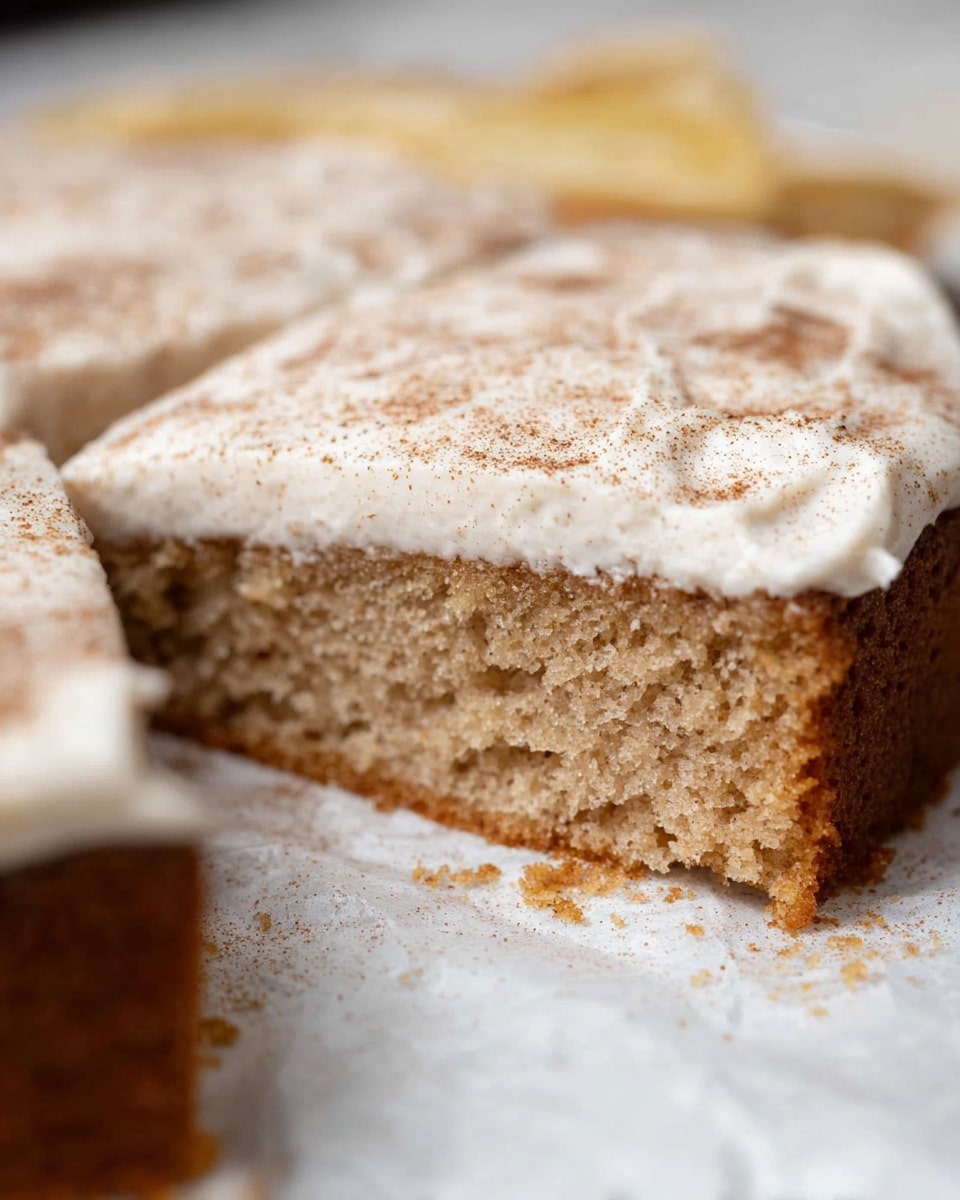A close-up view of a sliced, single-layer light brown cake with a soft and airy texture. The top layer is covered with a smooth white frosting sprinkled evenly with a fine dusting of brown cinnamon powder. The cake is presented on parchment paper, resting on a white marbled surface. The edges of the cake are clean and sharp, showing the moist crumb inside. Photo taken with an iphone --ar 4:5 --v 7