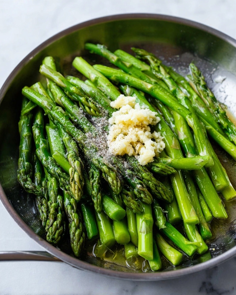 A close-up view of a metal pan filled with bright green asparagus stalks arranged in layers, topped in the center with a small pile of minced garlic, coarse salt, and ground black pepper, all sitting on a white marbled surface. The asparagus has a shiny, slightly wet texture, showing freshness and cooking preparation. photo taken with an iphone --ar 4:5 --v 7