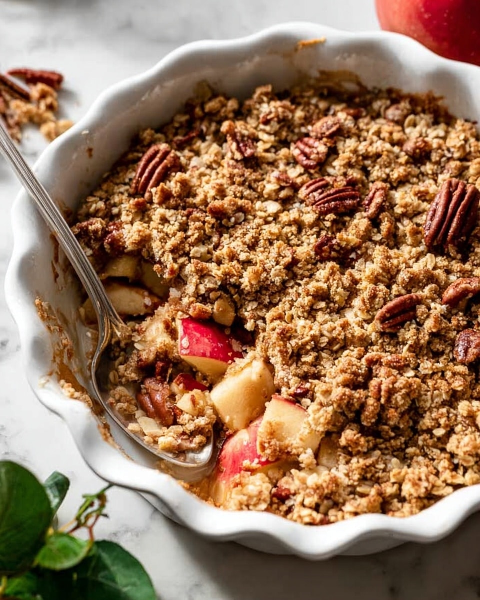 The image shows a white scalloped dish filled with apple crumble. The bottom layer has chunks of cooked apples with a light brown syrup, partially visible under the topping. The middle layer is a crunchy oat and pecan crumble, golden brown with a coarse texture. On top, there are three round scoops of vanilla ice cream, creamy white with small dark specks. A silver spoon rests inside the dish, scooping some of the crumble and apple mixture. Around the dish, fresh red apples with green leaves sit on a white marbled surface. photo taken with an iphone --ar 4:5 --v 7