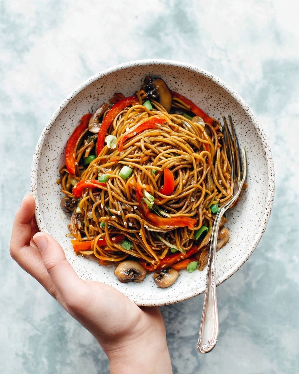 A woman's hand is holding a white, speckled bowl filled with a mound of brown noodles mixed with thin slices of red bell pepper, orange carrot strips, green onion pieces, and whole small brown mushrooms, with a silver fork resting on the side inside the bowl. The noodles are glossy and slightly twisted, layered on top of each other with the vegetables spread evenly throughout. The background has a white marbled texture. photo taken with an iphone --ar 4:5 --v 7