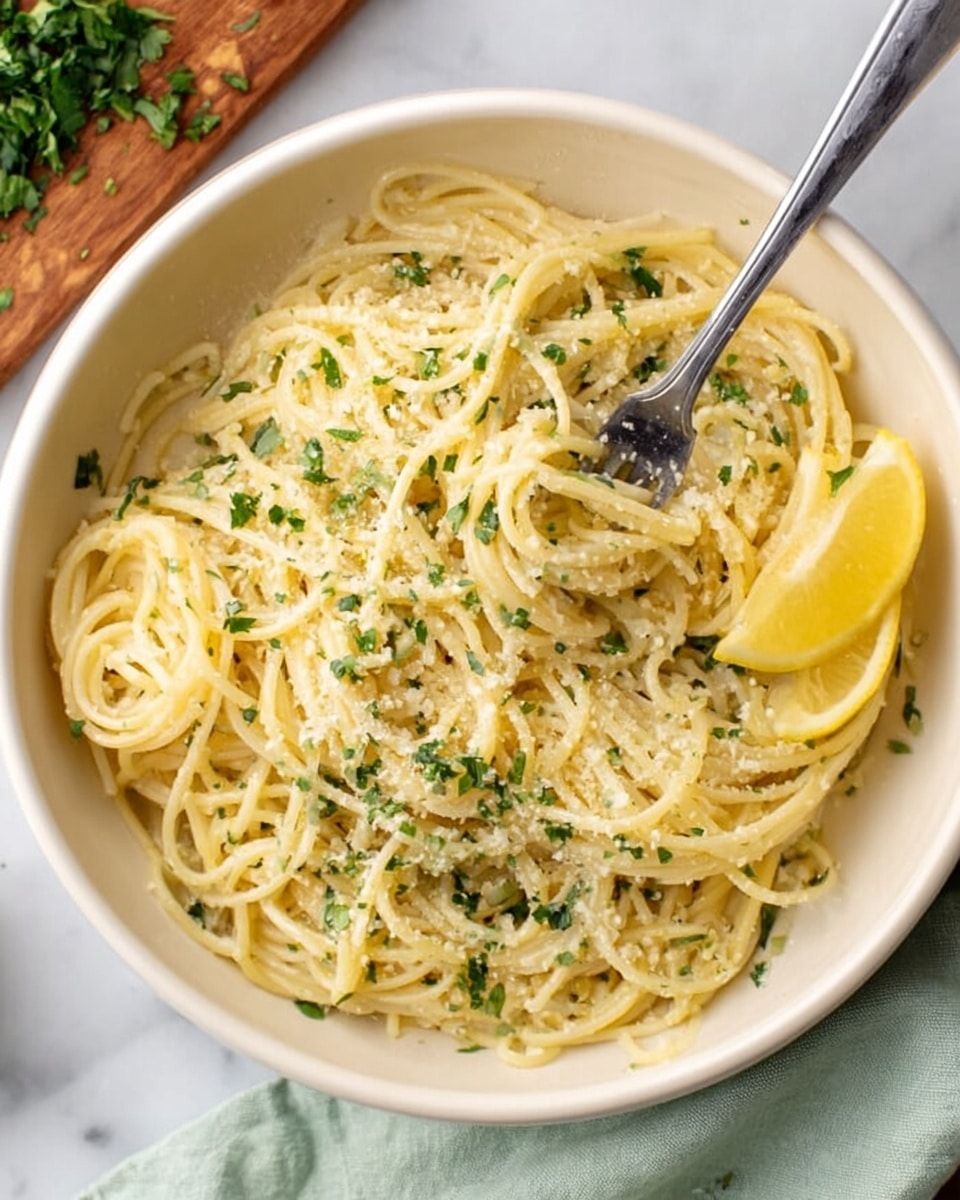 A white bowl filled with a single layer of cooked spaghetti noodles coated in a light sauce, sprinkled evenly with finely chopped green herbs and grated cheese. On the right edge of the bowl, there are two lemon wedges placed on top of the noodles. A silver fork rests inside the bowl, slightly twisting some noodles. The bowl is placed on a white marbled surface with a light green cloth nearby, and a wooden cutting board with fresh herbs is partially visible in the top left corner. Photo taken with an iphone --ar 4:5 --v 7
