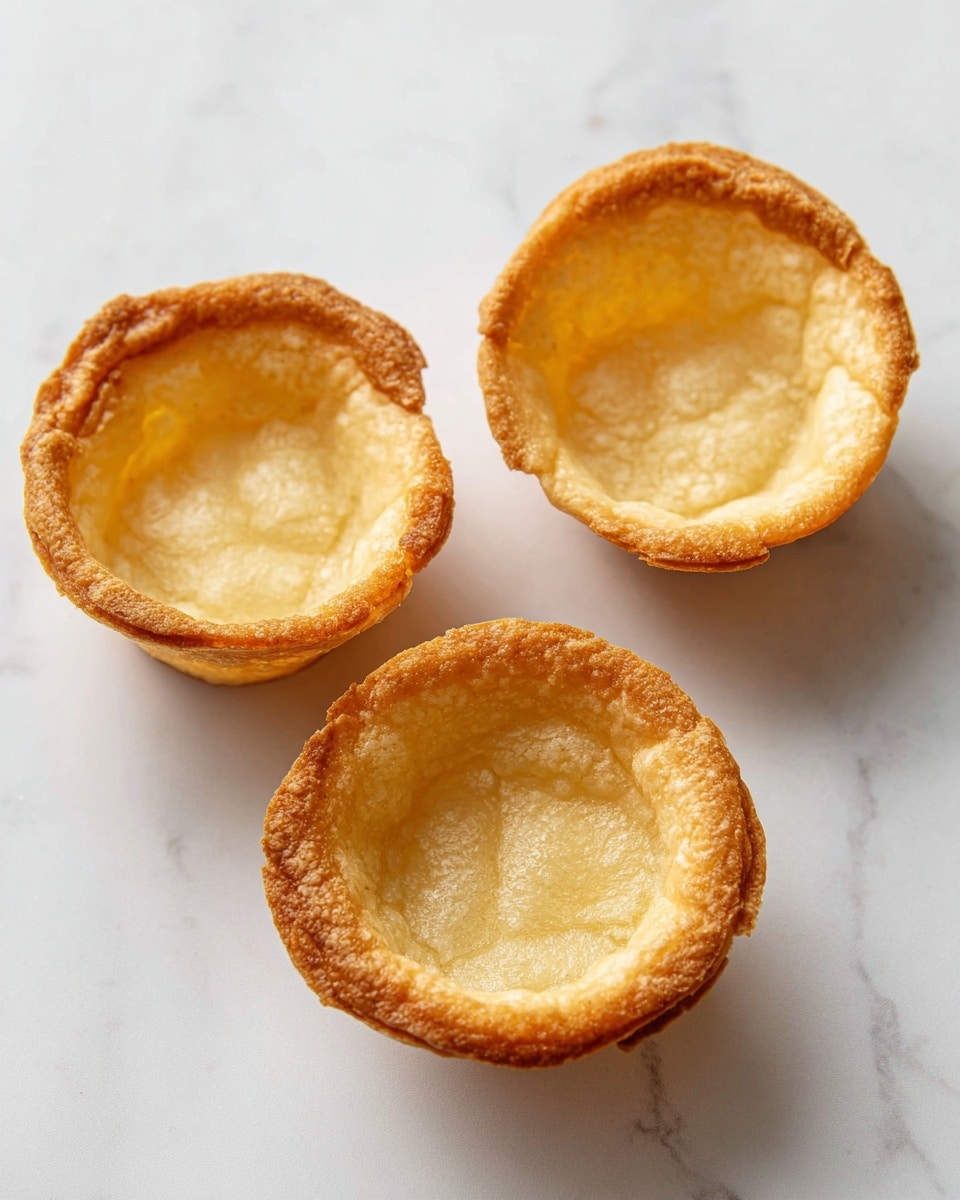 Three small round pastry shells are placed on a white marbled surface. Each shell has a golden-brown crispy outer edge and a lighter beige soft inside that looks smooth and slightly crumbly. The shells are arranged with one near the top and two closer together at the bottom, showing their hollow centers clearly. The lighting highlights the texture differences between the crispy outer rim and the soft inner base. Photo taken with an iphone --ar 4:5 --v 7