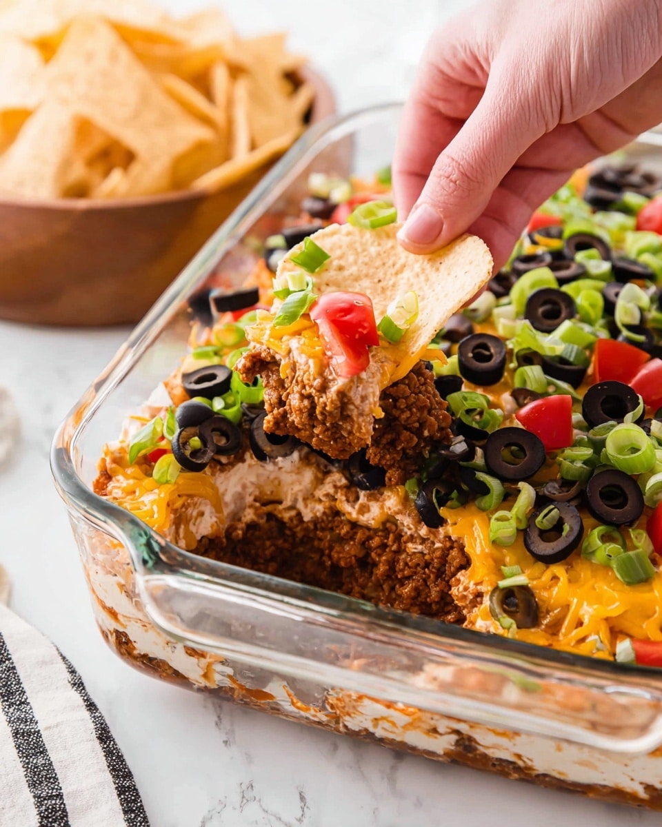 A clear square glass dish contains a layered dip with a base layer of light beige creamy spread, followed by a rich brown layer of ground meat sauce, and a generous middle layer of bright orange shredded cheese. The top layer is full of black olive slices scattered evenly with chopped bright green spring onions and small pieces of red tomato. A woman's hand is holding a round, beige chip scooping up a portion from the dish. The dish sits on a white marbled surface with a wooden bowl filled with similar beige chips partially visible at the bottom left and a white cloth with black stripes at the bottom right. Photo taken with an iphone --ar 4:5 --v 7