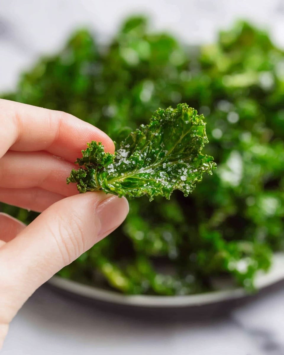 A close-up image of a woman's hand holding a single crisp kale chip, bright green with a slightly oily shine and salt crystals visible on its textured, curly edges. In the background, there is a shallow white plate filled with a large pile of similar crisp kale chips, all vibrant green with a crunchy texture, resting on a white marbled surface. The overall look is fresh and crunchy with natural green and white colors. photo taken with an iphone --ar 4:5 --v 7