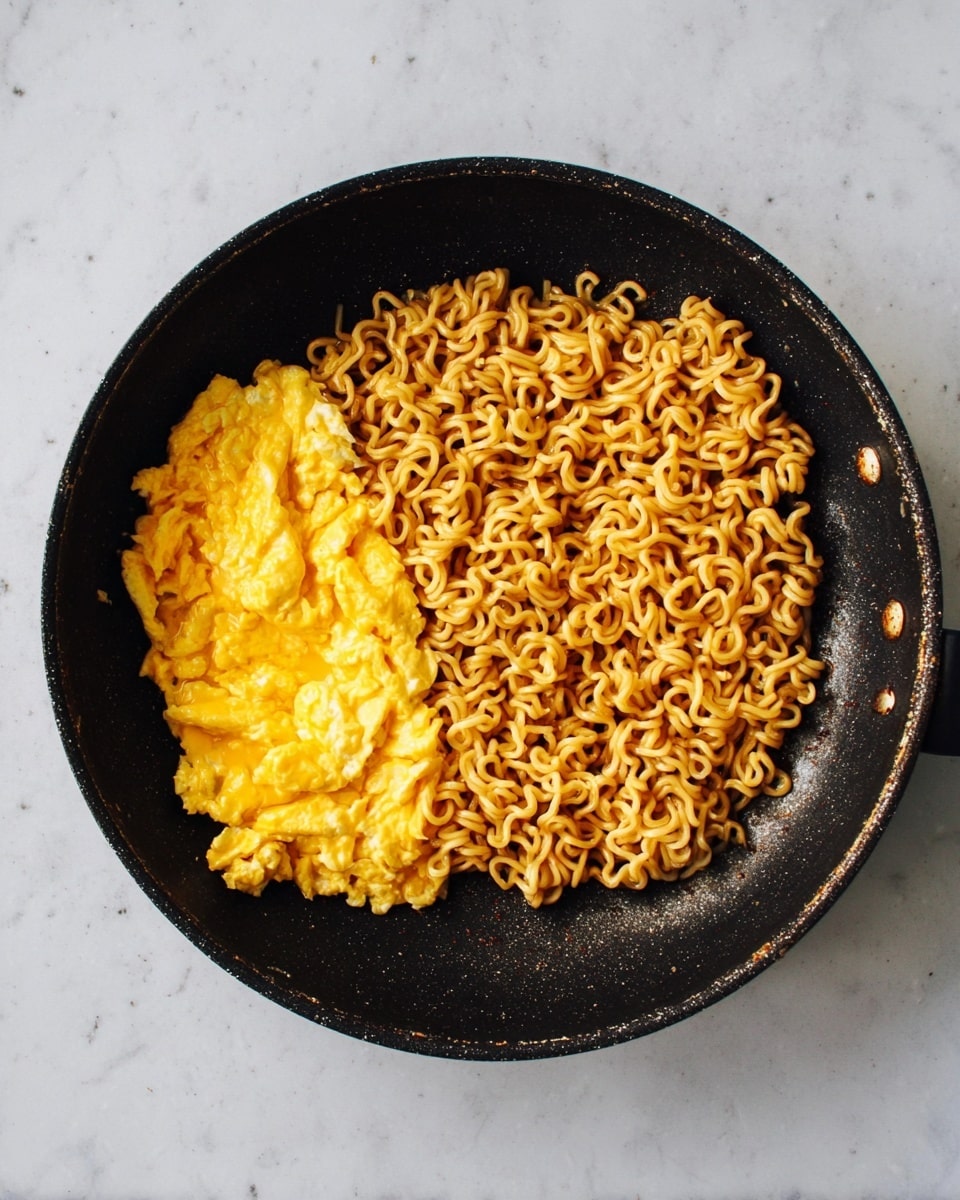 A black frying pan holds a single layer of cooked curly ramen noodles on the right side, golden brown with slight dark spots showing light crispness, while on the left side there is a folded layer of bright yellow scrambled eggs with a soft, slightly shiny texture. The pan sits on a white marbled surface, with no extra items around. Photo taken with an iphone --ar 4:5 --v 7