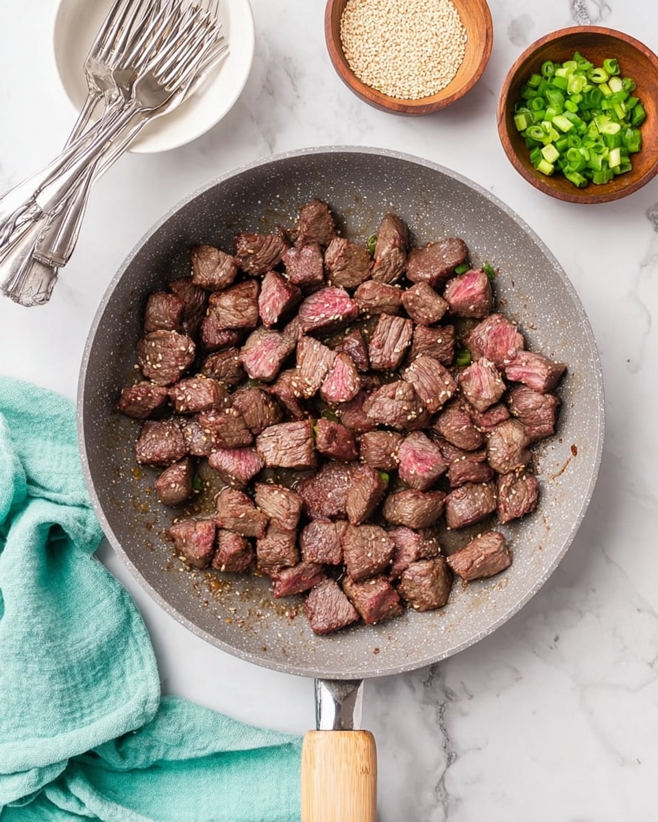 A gray frying pan filled with many small pieces of partly cooked beef, showing a mix of brown seared surfaces and pink centers, spread evenly across the pan's surface. The pan has a light wooden handle at the bottom. To the top left, there is a white bowl holding three forks with shiny metal handles. Above the pan, two small brown wooden bowls are placed on a white marbled surface; the left bowl contains pale sesame seeds, and the right bowl holds chopped green onions with white ends. A light turquoise cloth is partly visible near the bottom left corner. Photo taken with an iphone --ar 4:5 --v 7