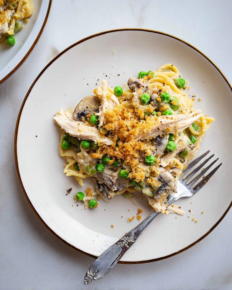 The image shows a close-up of a baked casserole in a round white dish with a golden brown, crispy breadcrumb topping that covers the surface evenly. Beneath the crumb layer, there is a creamy mix of white pasta pieces, green peas, light beige chunks of cooked chicken, and slices of brown mushrooms. A silver spoon rests inside the dish, scooping out some of the casserole, showing its soft, creamy texture and the mix of ingredients. The dish sits on a white marbled surface. photo taken with an iphone --ar 4:5 --v 7
