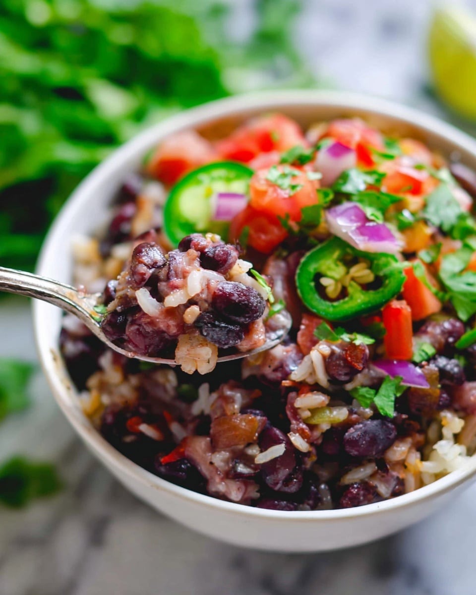 A white bowl filled with a colorful mix of black beans and rice as the base layer, showing a soft and slightly chunky texture with dark purples and browns. On one side, a fresh topping layer of bright red diced tomatoes, chopped green cilantro, small black beans, thin slices of green jalapeño with visible seeds, and bits of red onion adds vibrant red, green, purple, and white colors. A silver spoon lifts a scoop of the bean and rice mix, showcasing the moist and mixed texture. The bowl sits on a white marbled surface, with a blurred background of green leafy vegetables. Photo taken with an iphone --ar 4:5 --v 7
