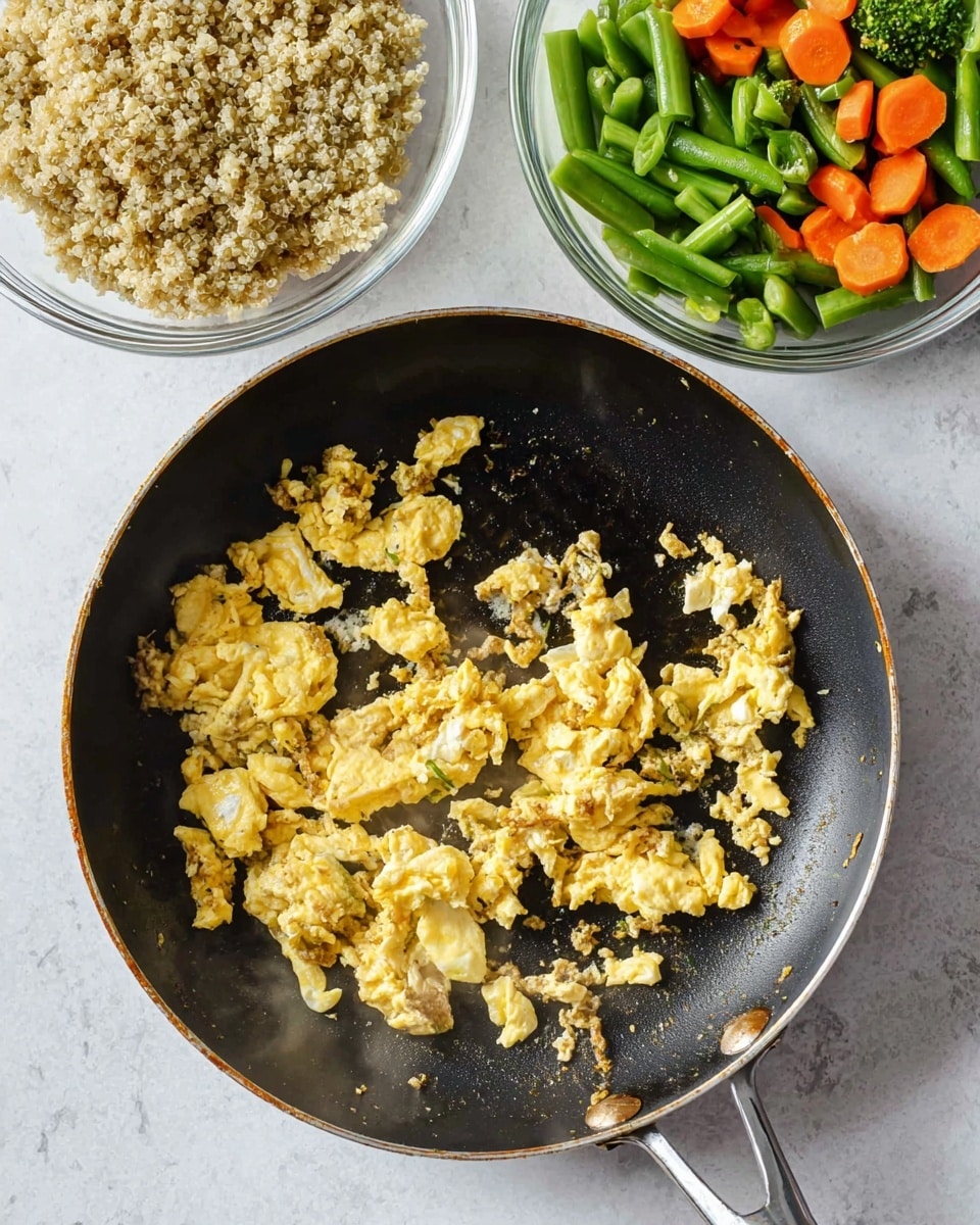 A black frying pan with a silver handle holds scrambled eggs cooking in it, showing a mix of fluffy, slightly browned, and soft white egg pieces scattered unevenly on the pan's surface, with some steam rising. Above the pan, two clear glass bowls sit side by side on a white marbled surface. The bowl on the left contains cooked quinoa with light beige and some dark grains mixed in, giving it a fluffy, grainy texture. The bowl on the right holds frozen mixed vegetables, showing bright green broccoli, green beans, celery, and orange carrot pieces with a slight frost covering. Photo taken with an iphone --ar 4:5 --v 7