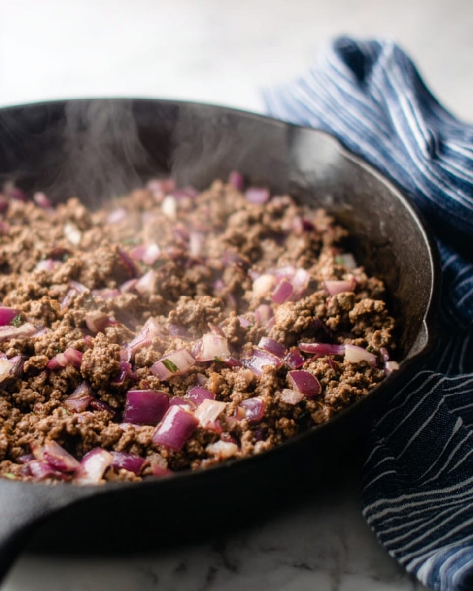 The image shows a black cast iron pan filled with browned ground meat mixed with small pieces of cooked red and white onions, creating a mix of brown and purple colors with a slightly chunky texture. The pan sits on a white marbled surface, and behind it, there is a blue and white striped kitchen towel slightly out of focus. Steam rises softly from the meat, indicating it is hot and freshly cooked. Photo taken with an iphone --ar 4:5 --v 7