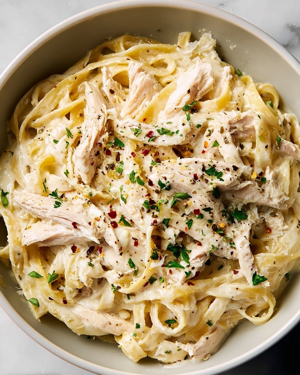 The image shows a white bowl filled with creamy fettuccine pasta mixed with slices of cooked white meat chicken. The pasta is pale yellow and coated well with a smooth, creamy white sauce. The dish is topped with small green parsley pieces, light brown ground black pepper, and tiny red flakes that add color contrast. The overall texture looks rich and hearty, with the pasta strands and chicken pieces intertwined. The bowl sits on a white marbled surface. photo taken with an iphone --ar 4:5 --v 7