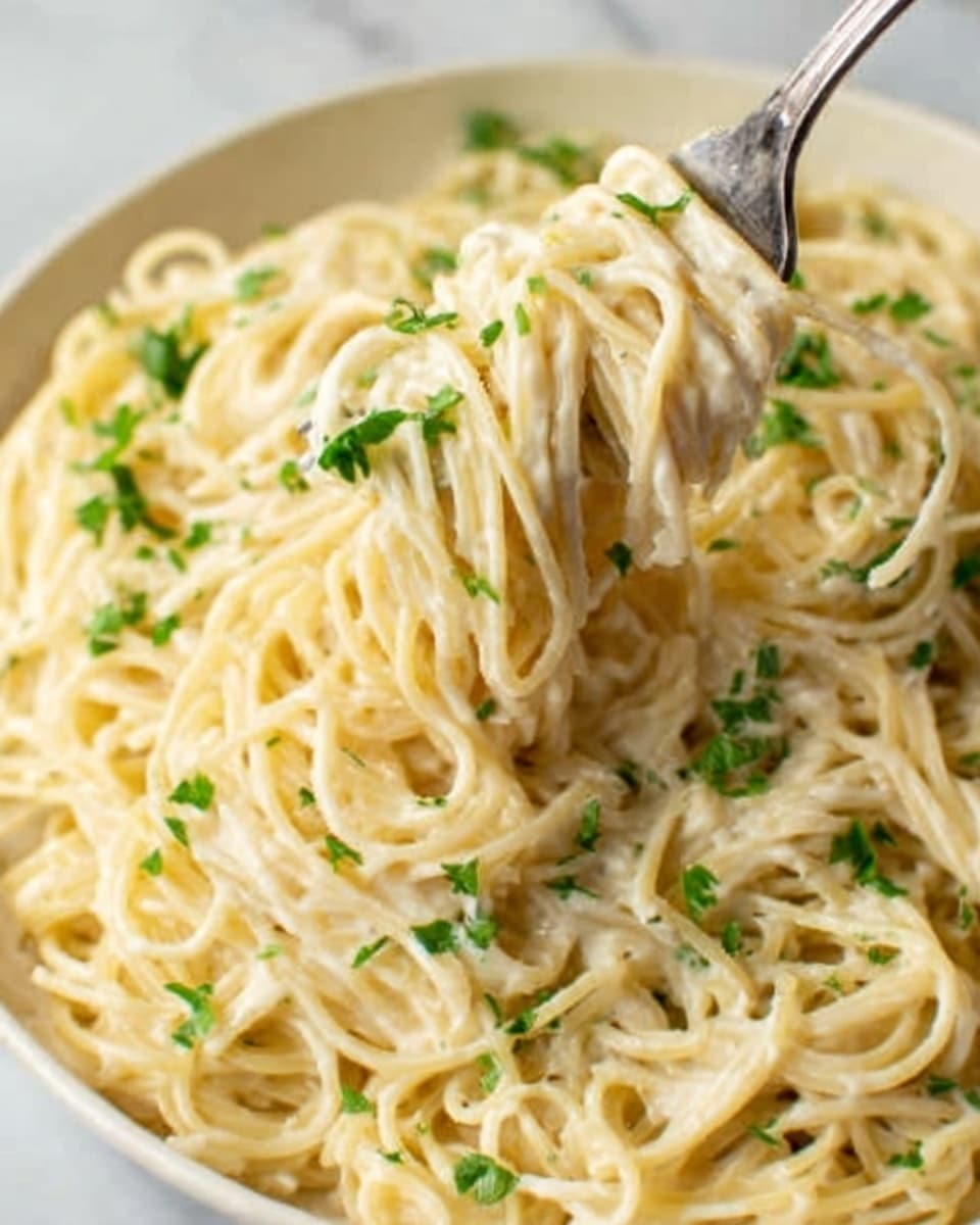 The image shows a close-up of a creamy pasta dish with thin spaghetti noodles coated evenly in a smooth, light beige sauce. The pasta is mixed well and sprinkled with small green parsley pieces on top, adding a fresh color contrast. A silver fork is lifting a small portion of the pasta, twisting the noodles gently. The dish is served on a white plate, placed on a white marbled surface. photo taken with an iphone --ar 4:5 --v 7